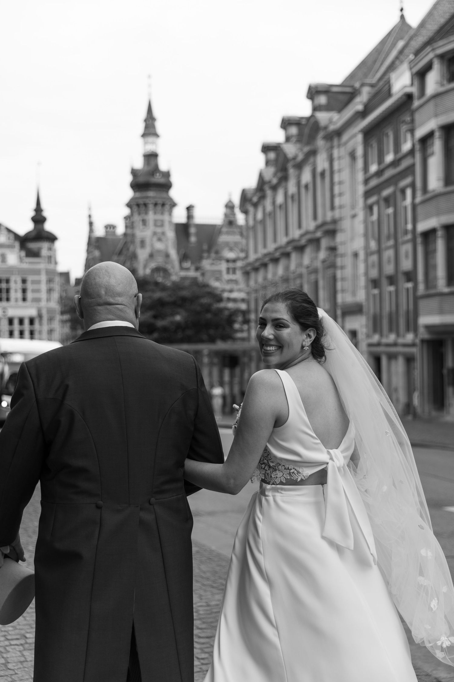 Black and white photo of bride and groom walking through historic European city square on wedding day.