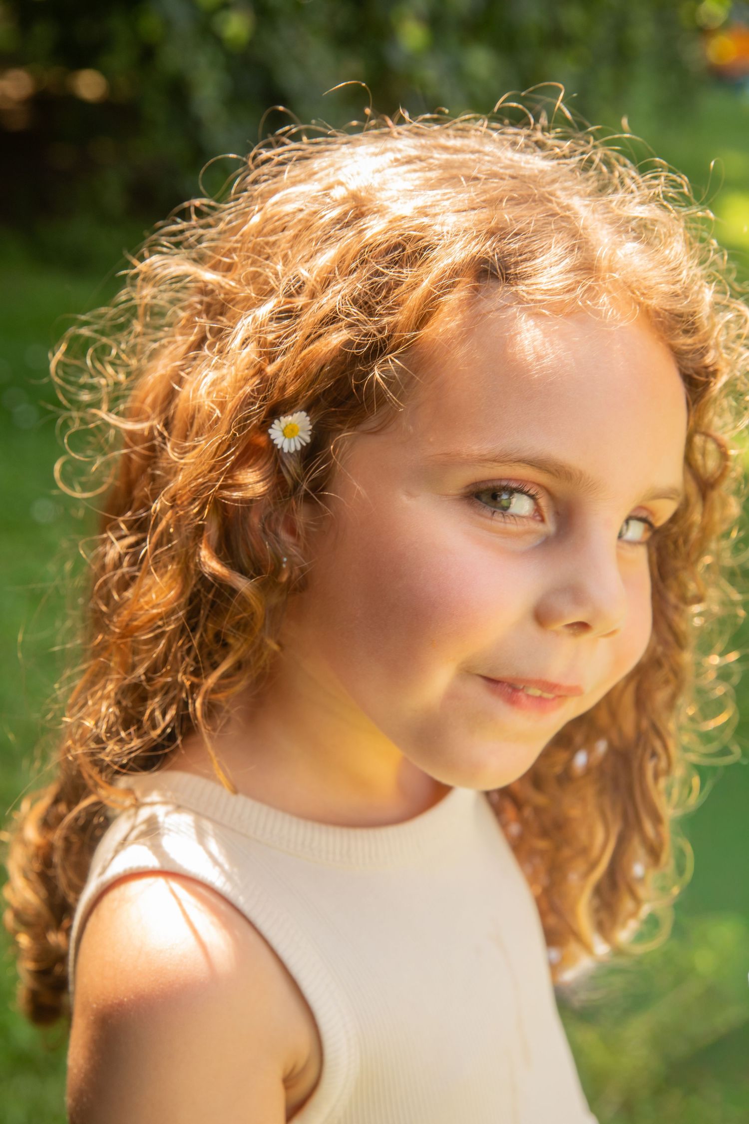 Young girl with curly red hair and a daisy clip smiles softly outdoors in warm sunlight.