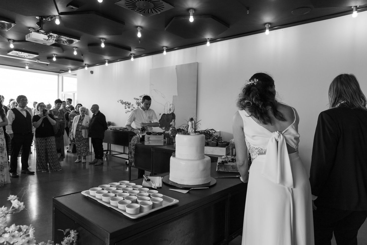 Black and white wedding reception photo showing bride at cake table with guests gathered in elegant venue.