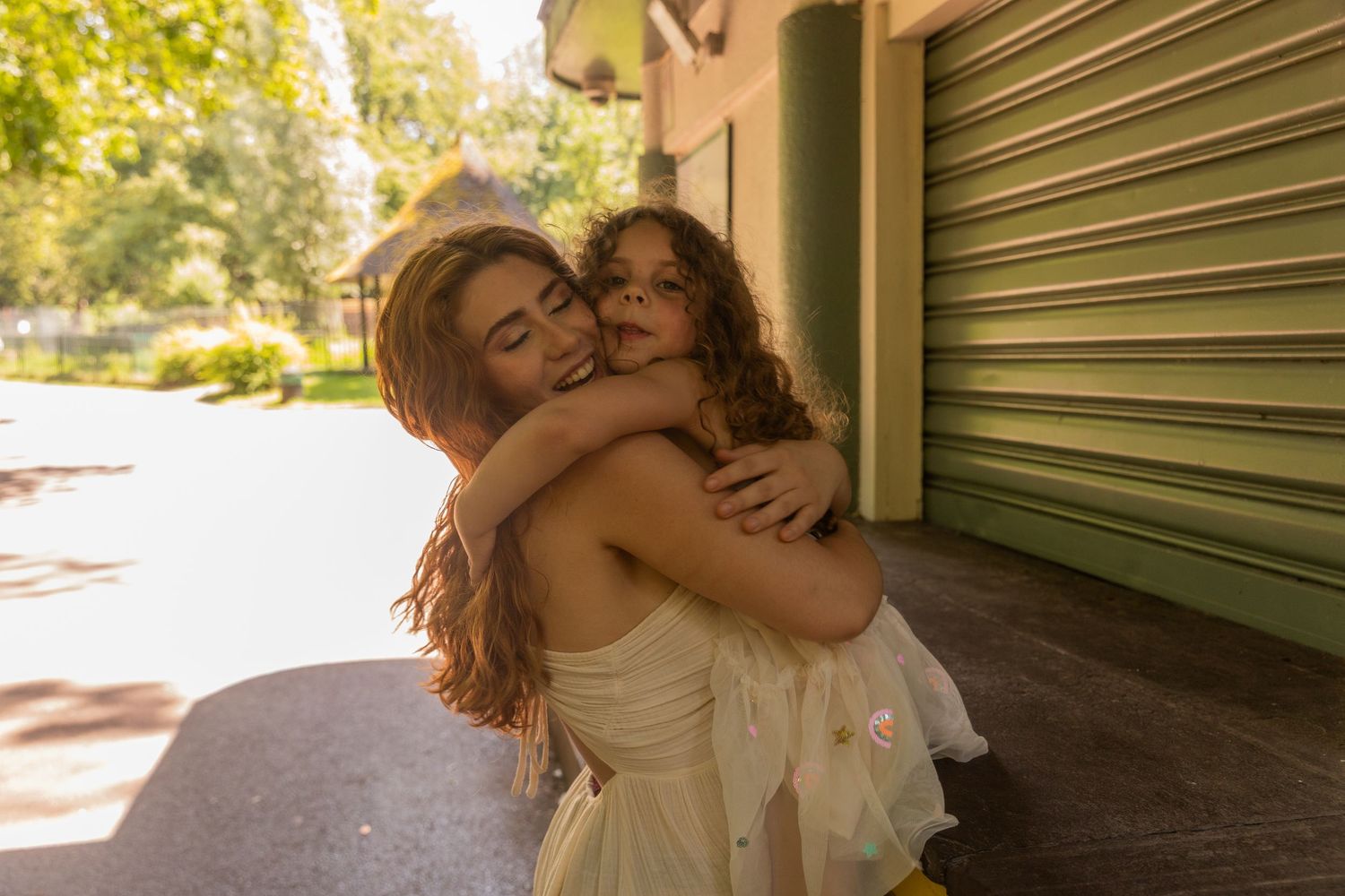 Two girls in light dresses share a warm hug outdoors on a sunny day near a green building.