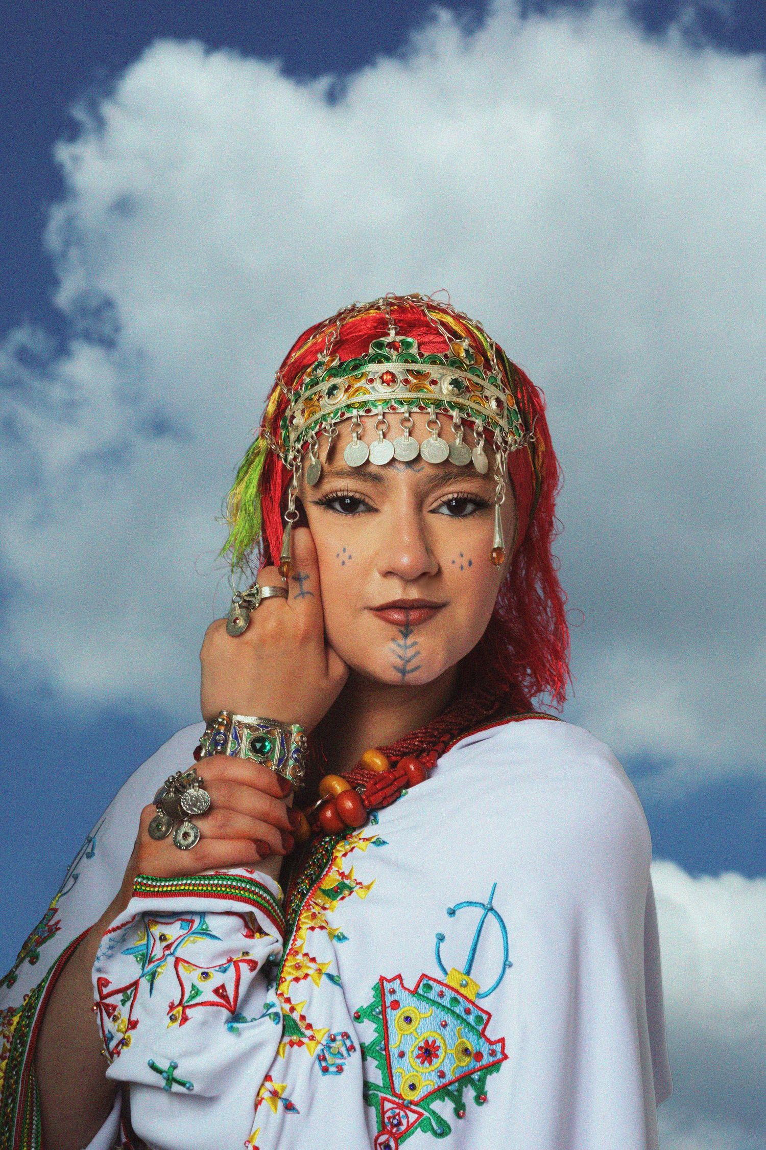 Woman in traditional Berber attire with ornate headdress, silver jewelry, and face markings poses against a cloudy sky.