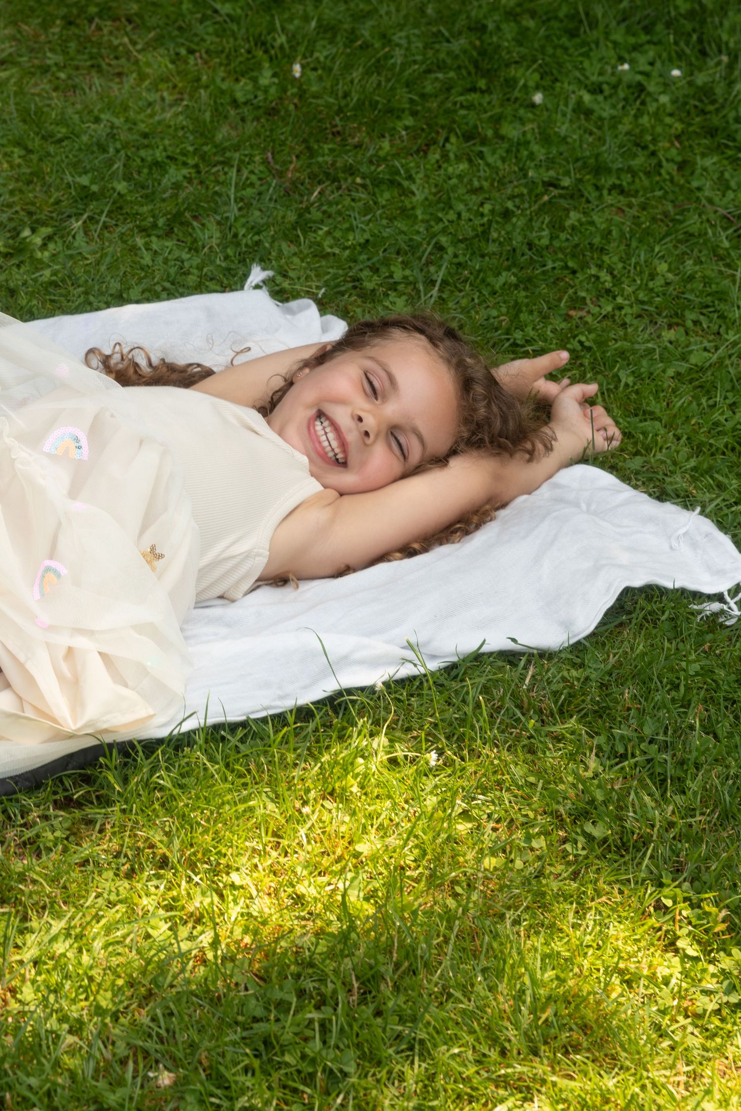 Happy young girl lying on a white blanket in lush green grass, smiling joyfully on a sunny day.