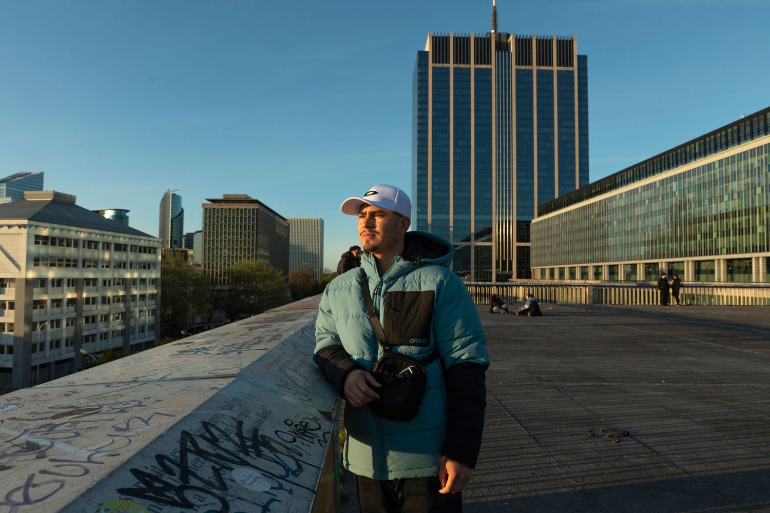 A young man in a blue jacket and white cap stands on a rooftop with a city skyline and modern buildings behind him.
