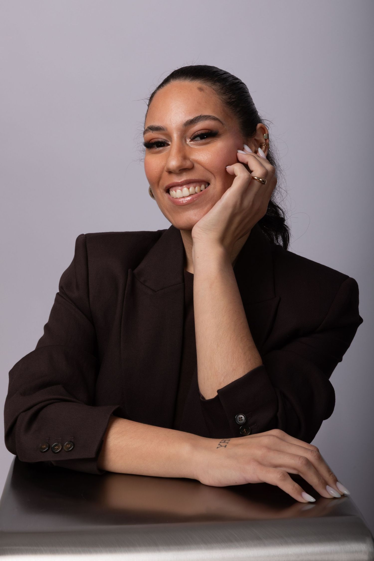 Smiling woman in dark blazer rests chin on hand at table, wearing rings and small wrist tattoo.