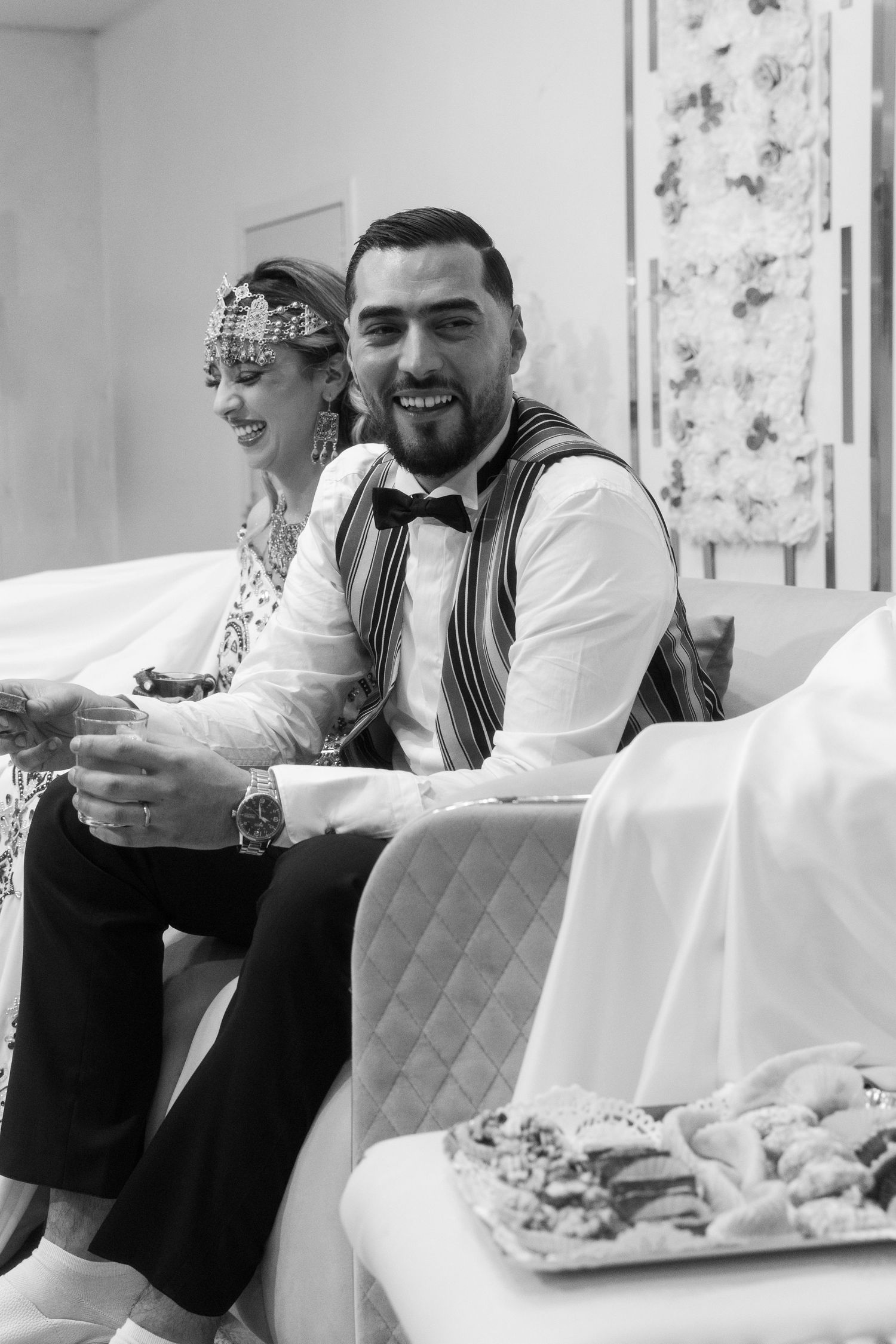 Laughing groom in vest and bow tie sits beside bride in traditional attire at wedding celebration.