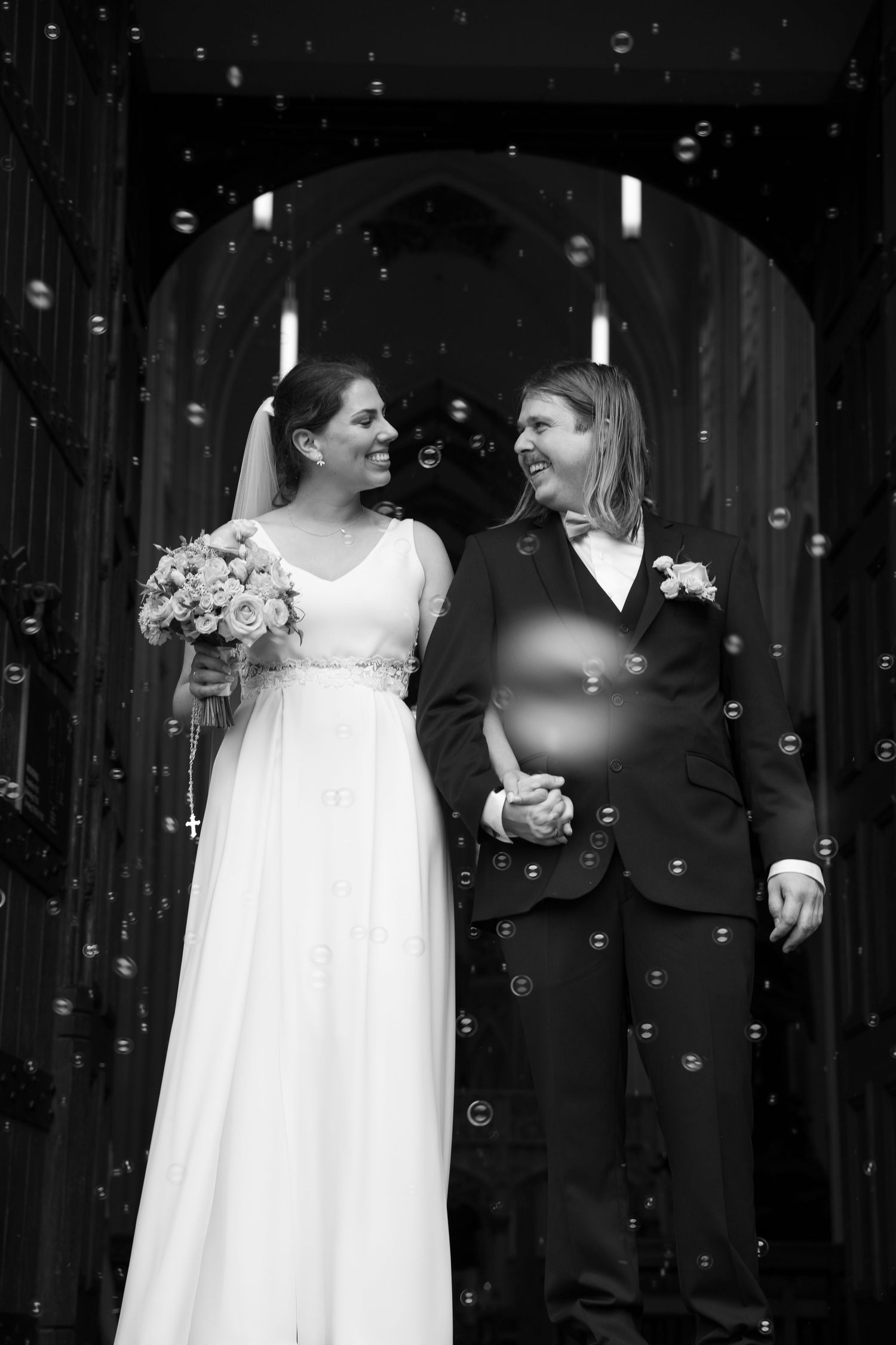 Black and white photo of two brides holding hands and laughing surrounded by bubbles outside a church.