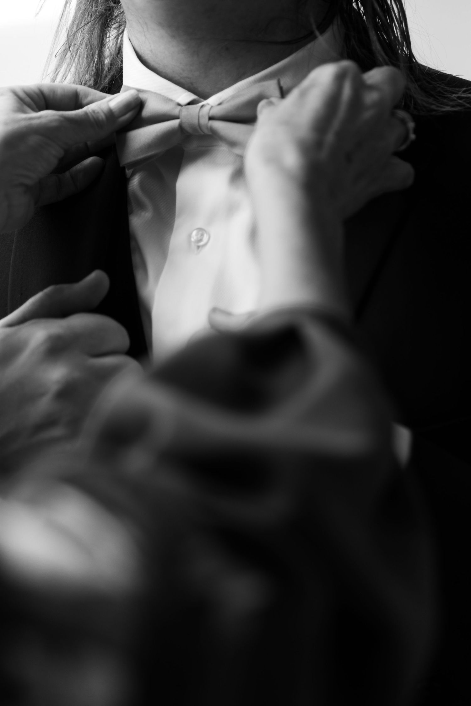 Black and white photo of hands adjusting a bow tie on a person wearing a formal suit and white dress shirt.