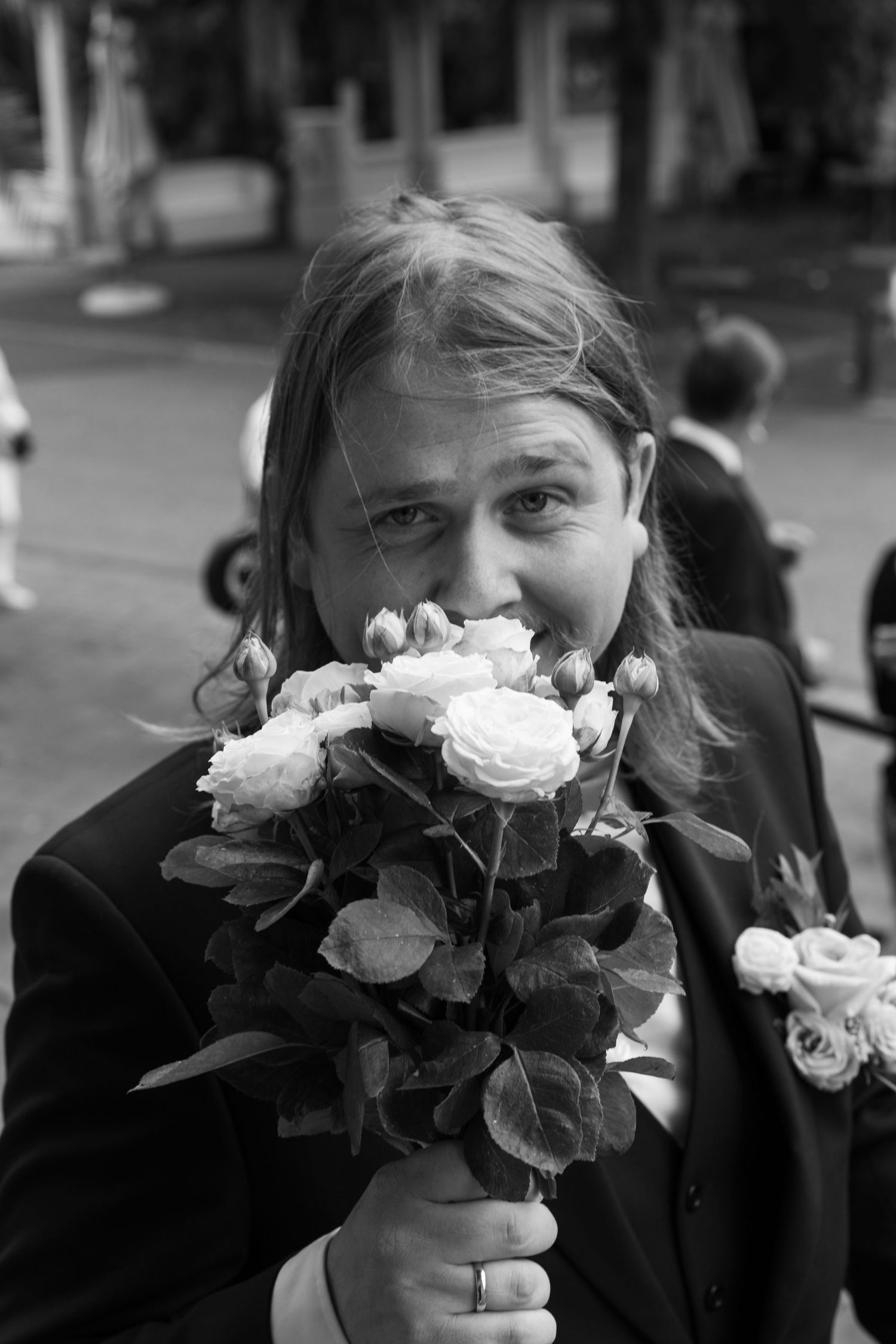 Black and white photo of a long-haired man in a suit smelling a bouquet of roses, wearing a ring.