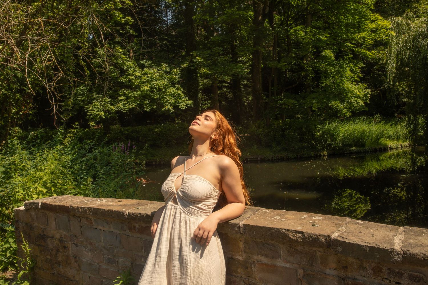 A woman in a white dress stands on a stone bridge, tilting her head back in sunlight, surrounded by lush green trees.