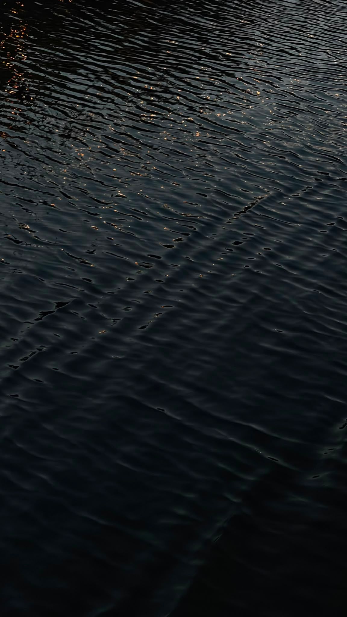 Dark rippling water surface with gentle waves reflecting dim light, creating a serene and moody aquatic texture pattern.