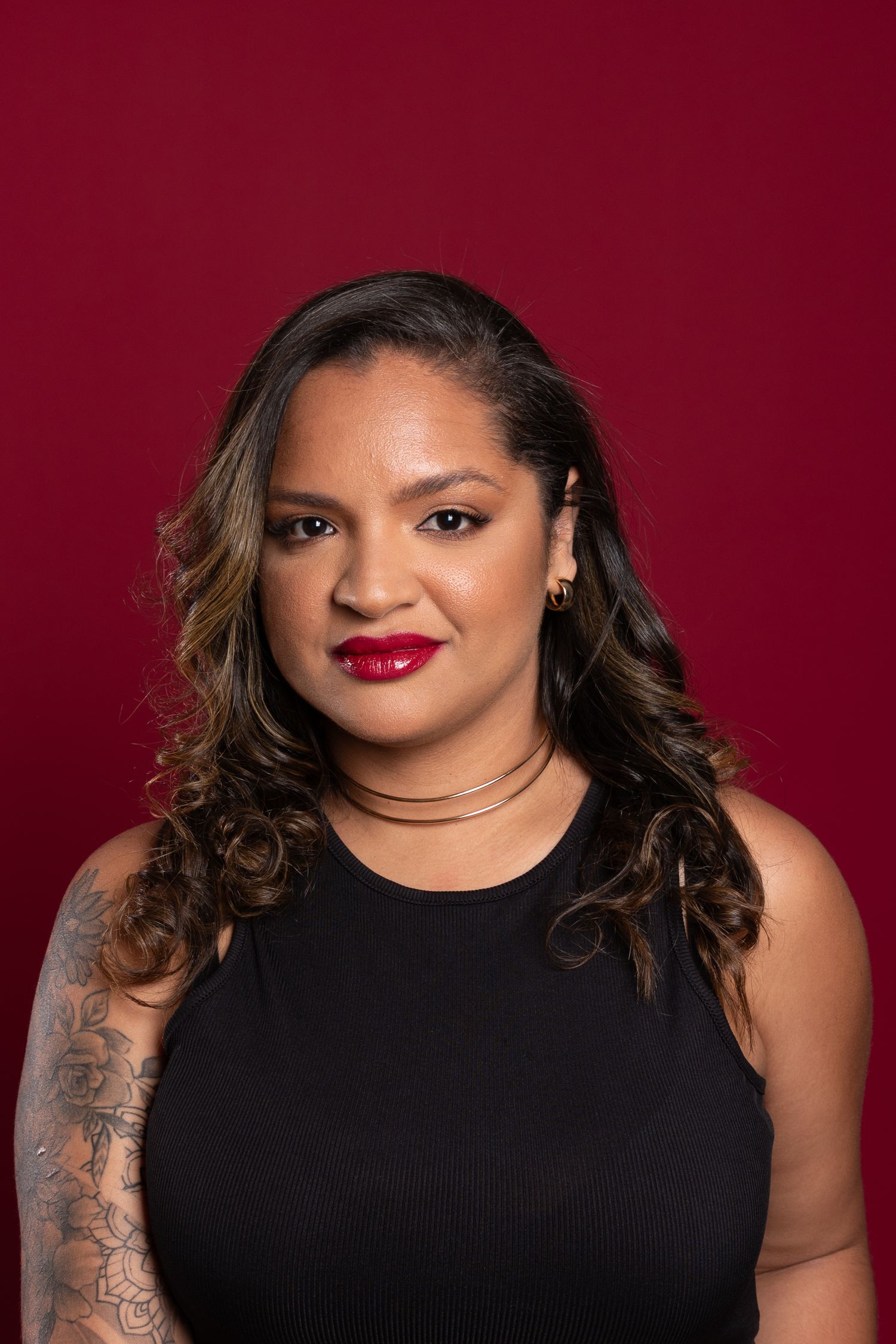 Woman with curly hair, red lipstick, gold necklace, floral arm tattoo, wearing black sleeveless top against red background.