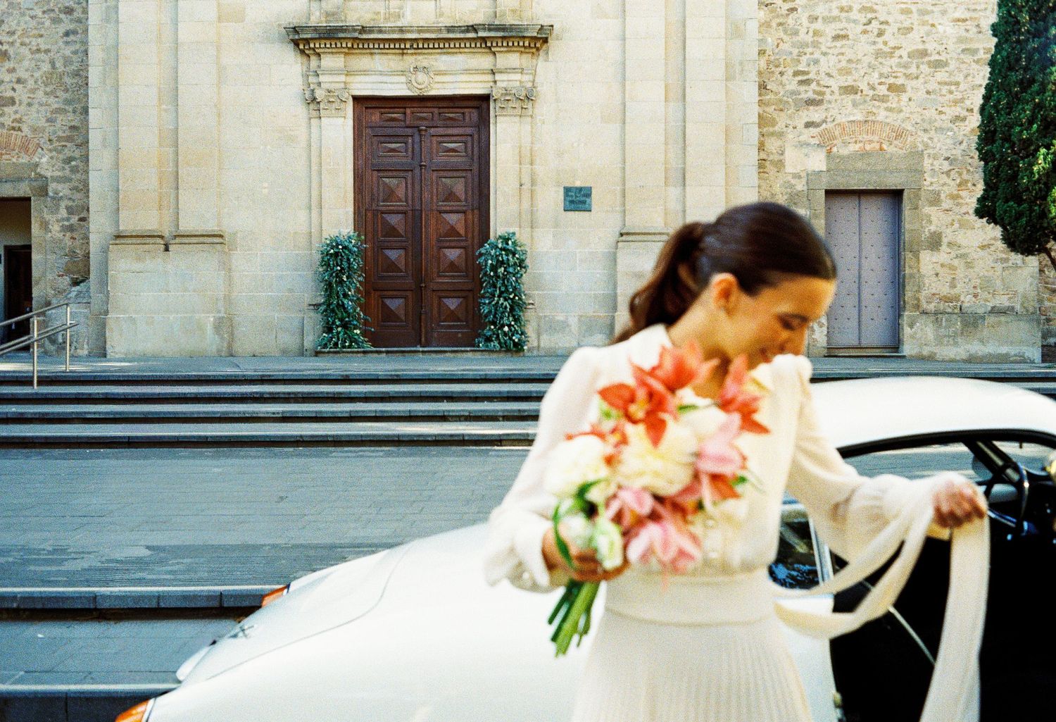 Bride in white dress holding a colorful bouquet of flowers beside a white car outside a historic church.