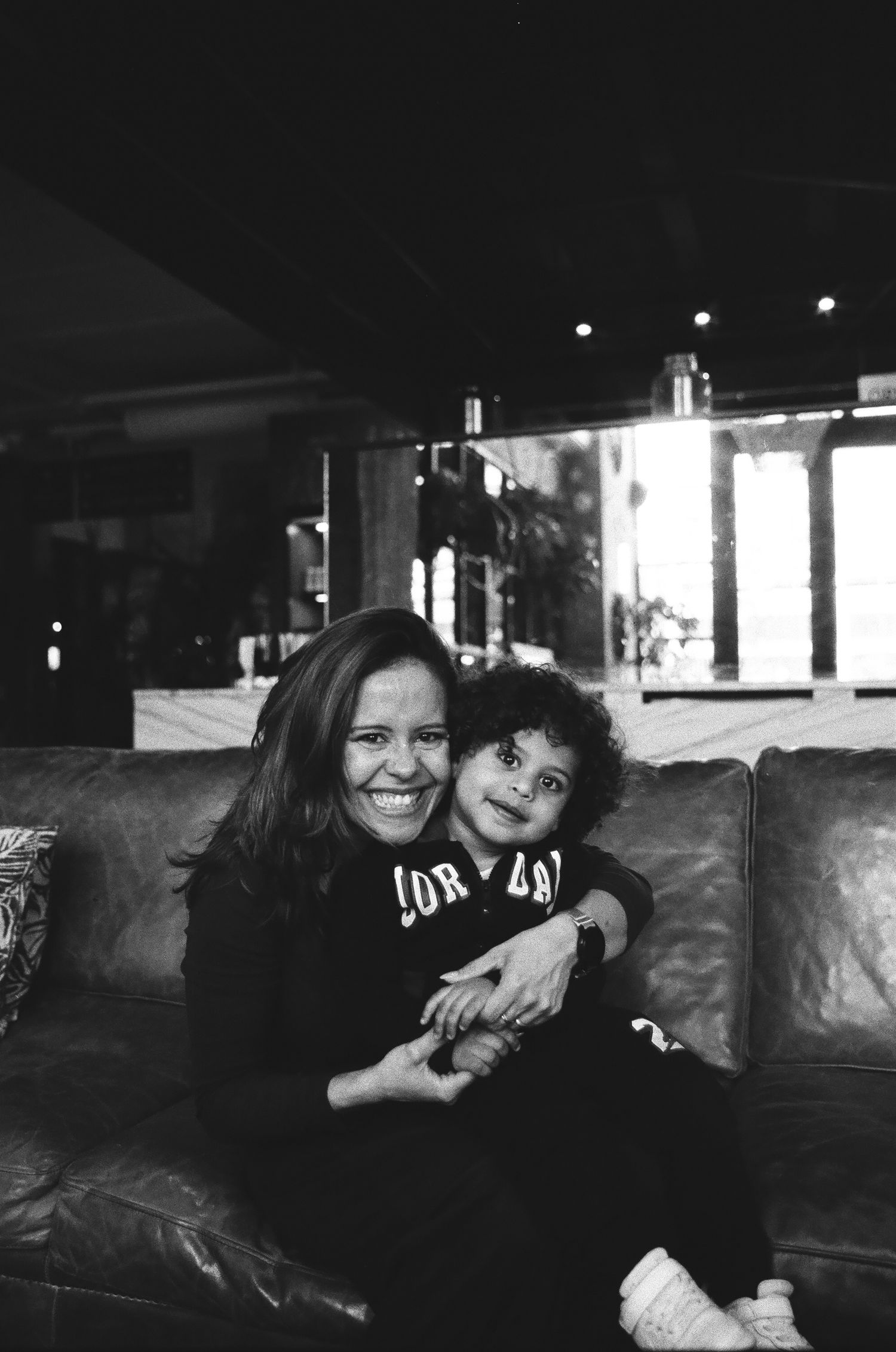 Black and white photo of a smiling woman hugging a young child on a leather couch indoors.