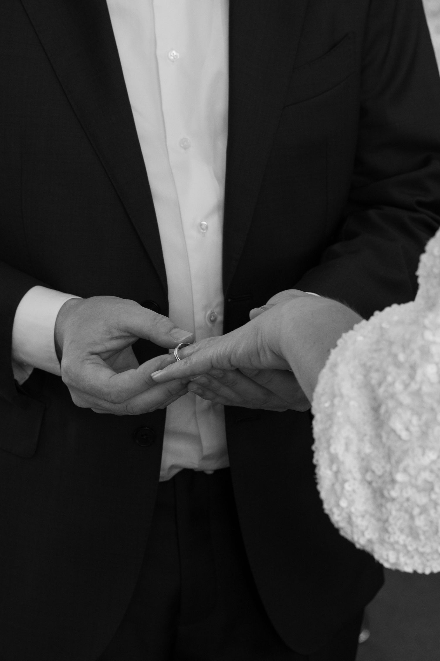 Black and white photo of groom placing wedding ring on bride's finger during ceremony.