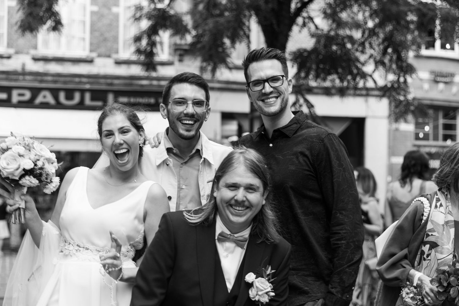 Black and white wedding photo of smiling bride and groom with two guests outdoors near a building.