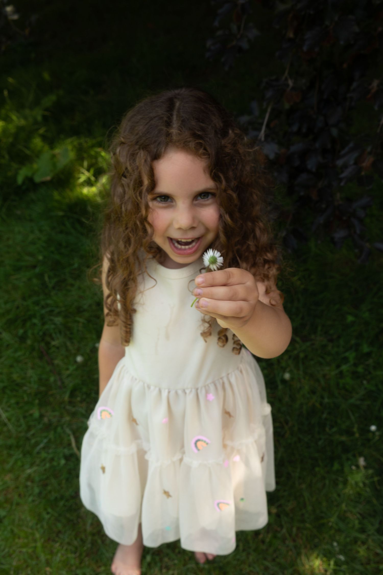 A smiling young girl with curly hair in a white dress holds a daisy flower outdoors in a lush green garden.