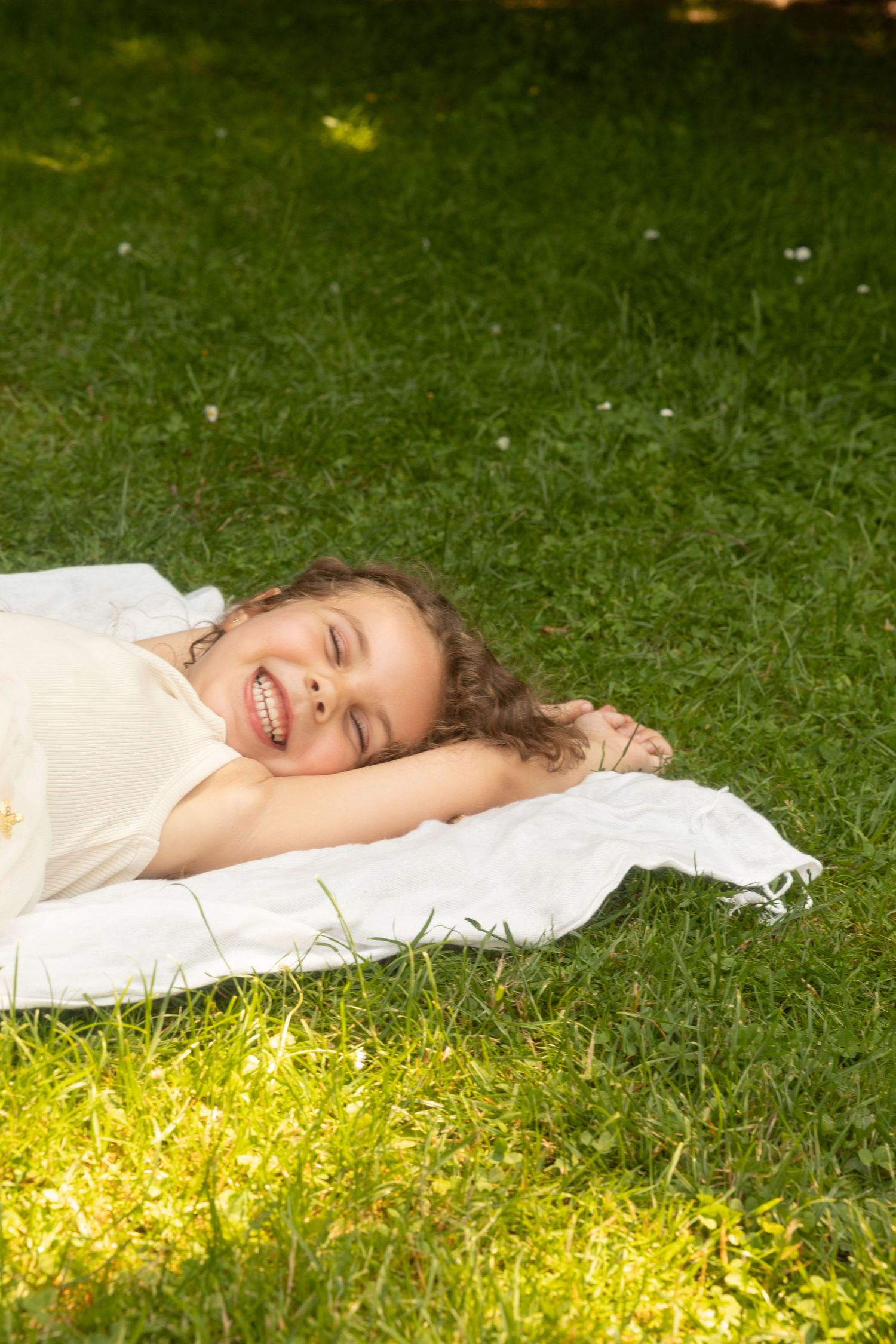 A happy young girl with curly hair lying on a white blanket in lush green grass, laughing joyfully outdoors.
