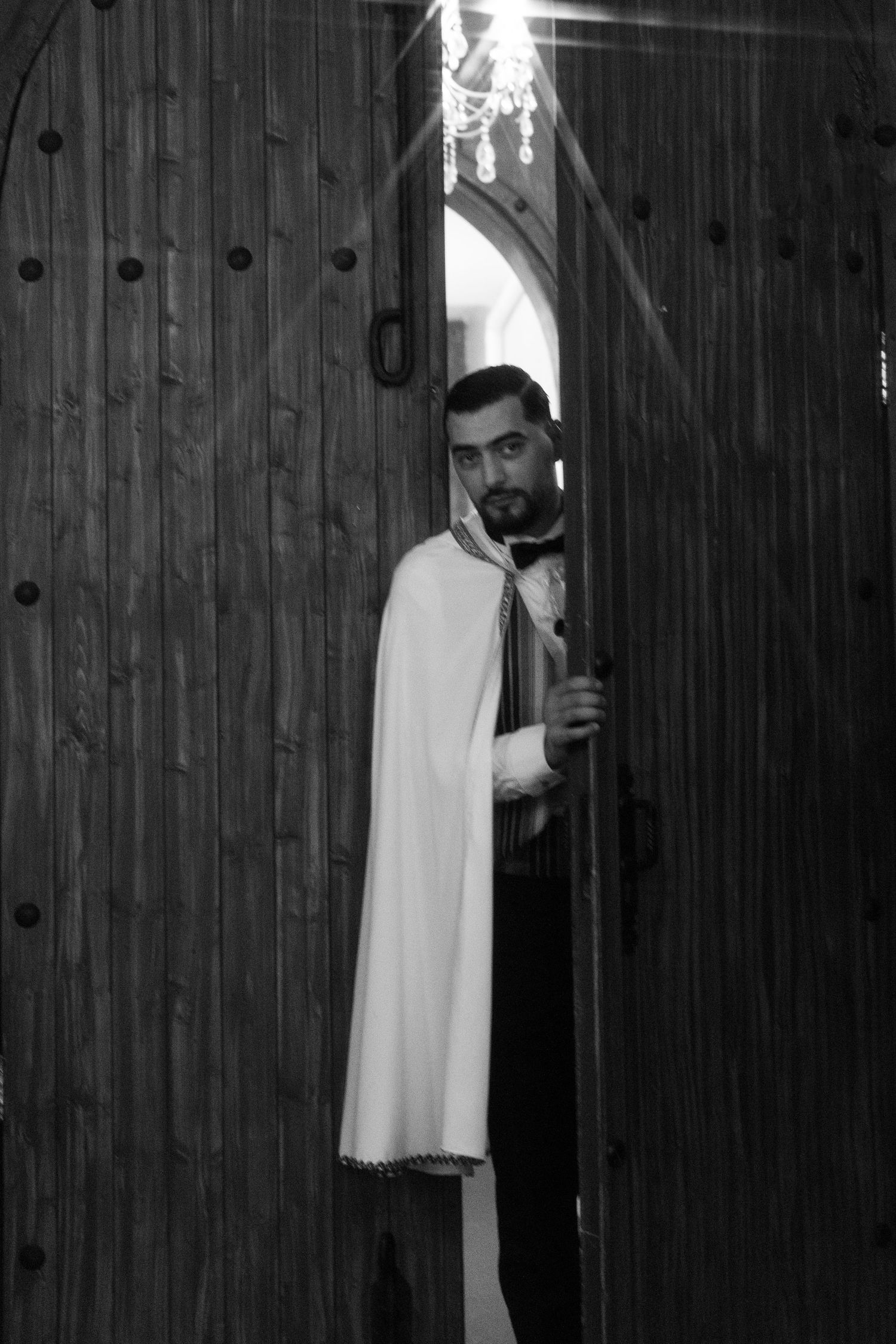 Black and white photo of priest peering through large wooden church doors before wedding ceremony.