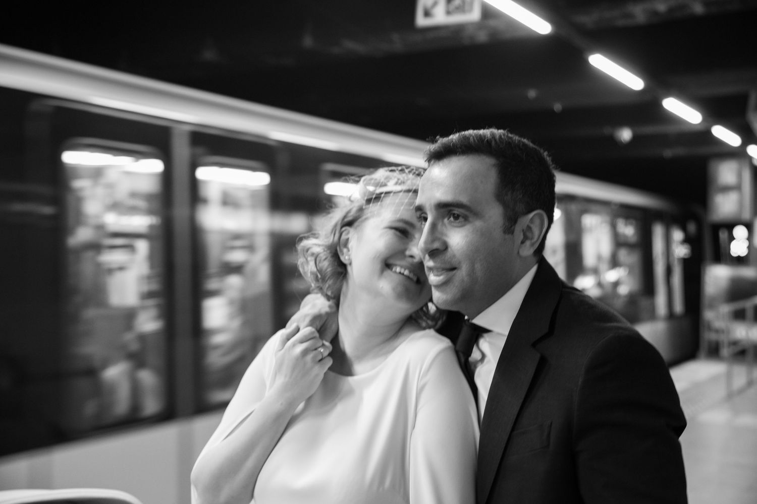 Black and white photo of a newlywed couple embracing at a subway station, bride in white dress, groom in suit.
