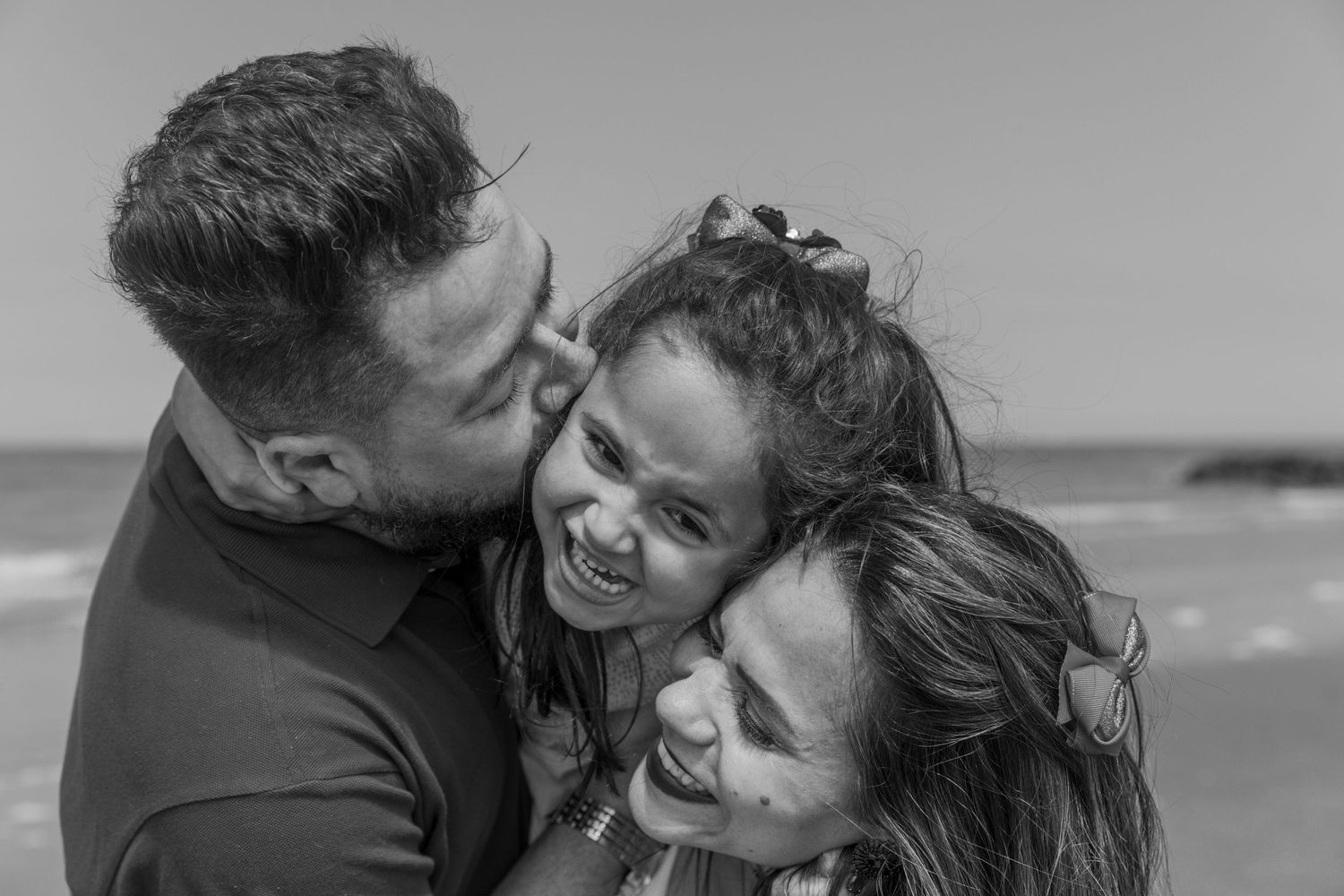 Black and white beach photo of a man and woman hugging and laughing with their young daughter. photography brussels