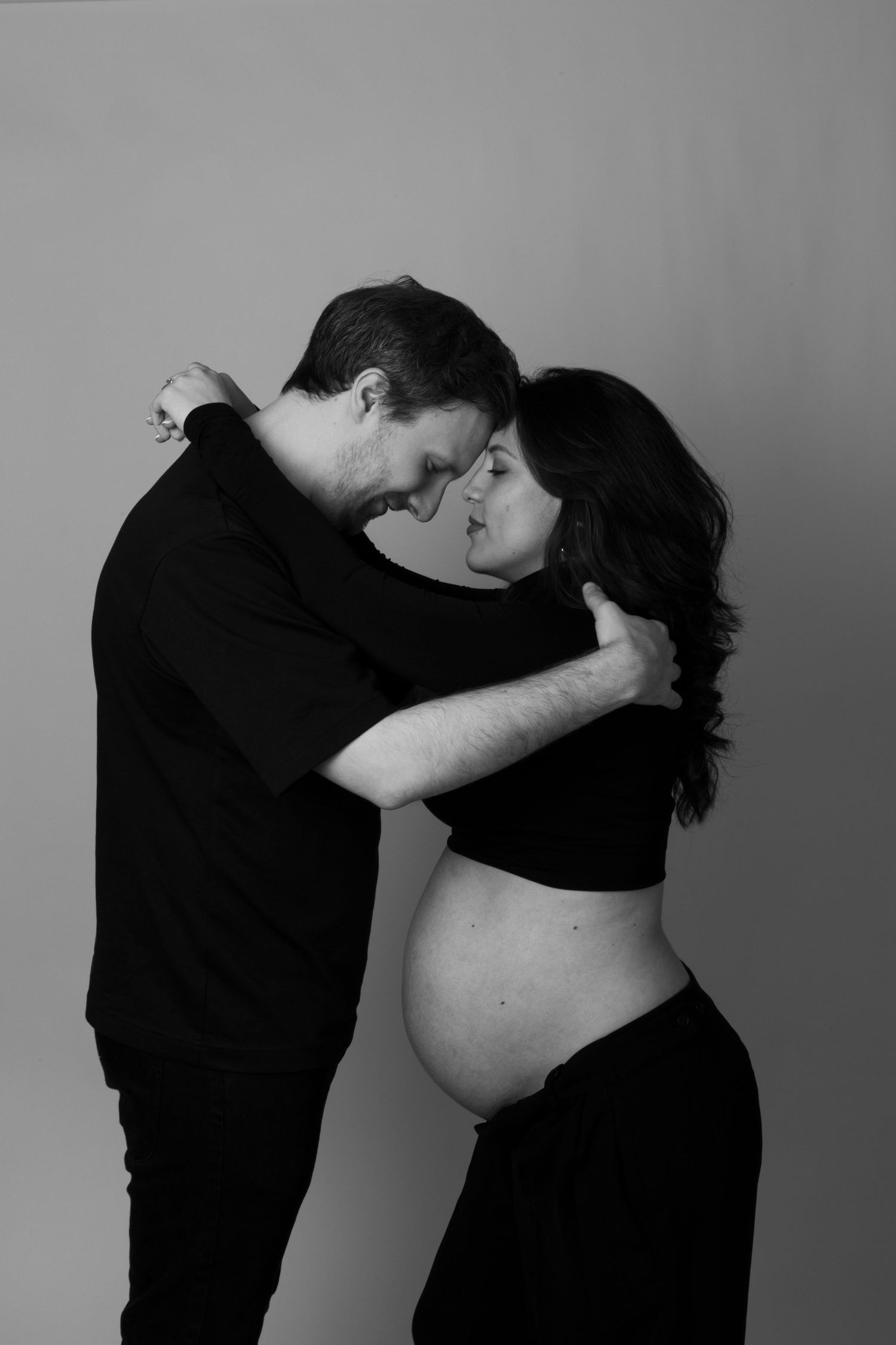 Black and white maternity photo of a couple embracing, showing pregnant belly against a neutral background. photography brussels
