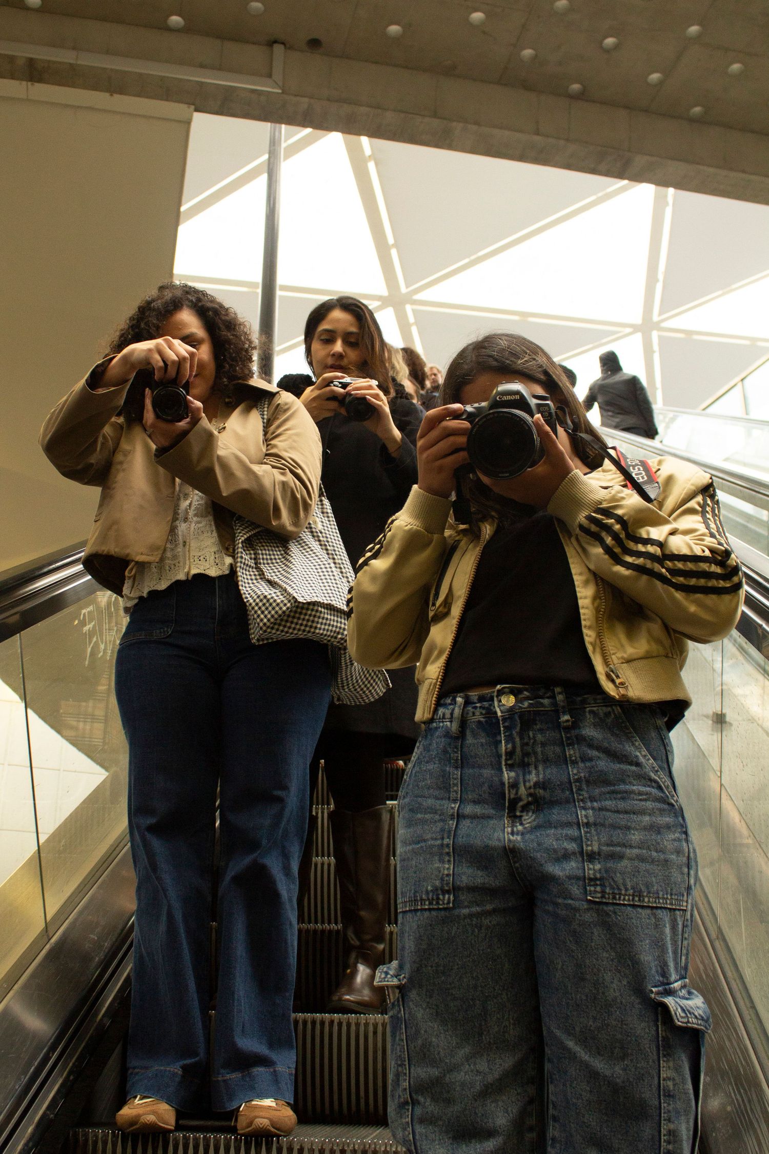 Three young women on an escalator holding cameras up to photograph, wearing casual jackets and jeans indoors.