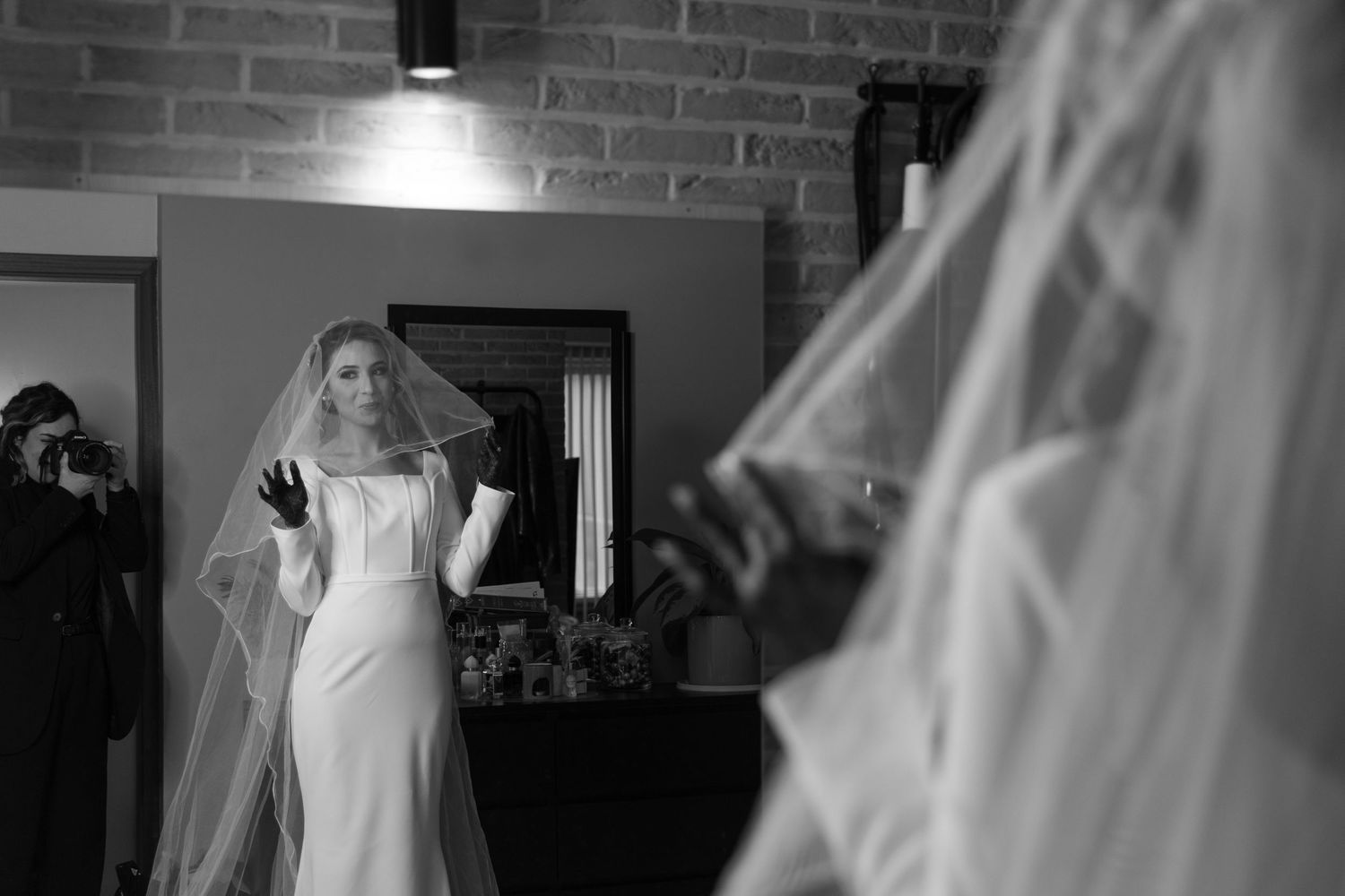 Bride in white wedding dress and veil admires her reflection in a mirror in black and white photo.