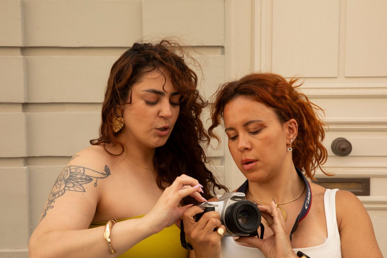 Two women examining photos on a camera together outdoors near a building entrance.