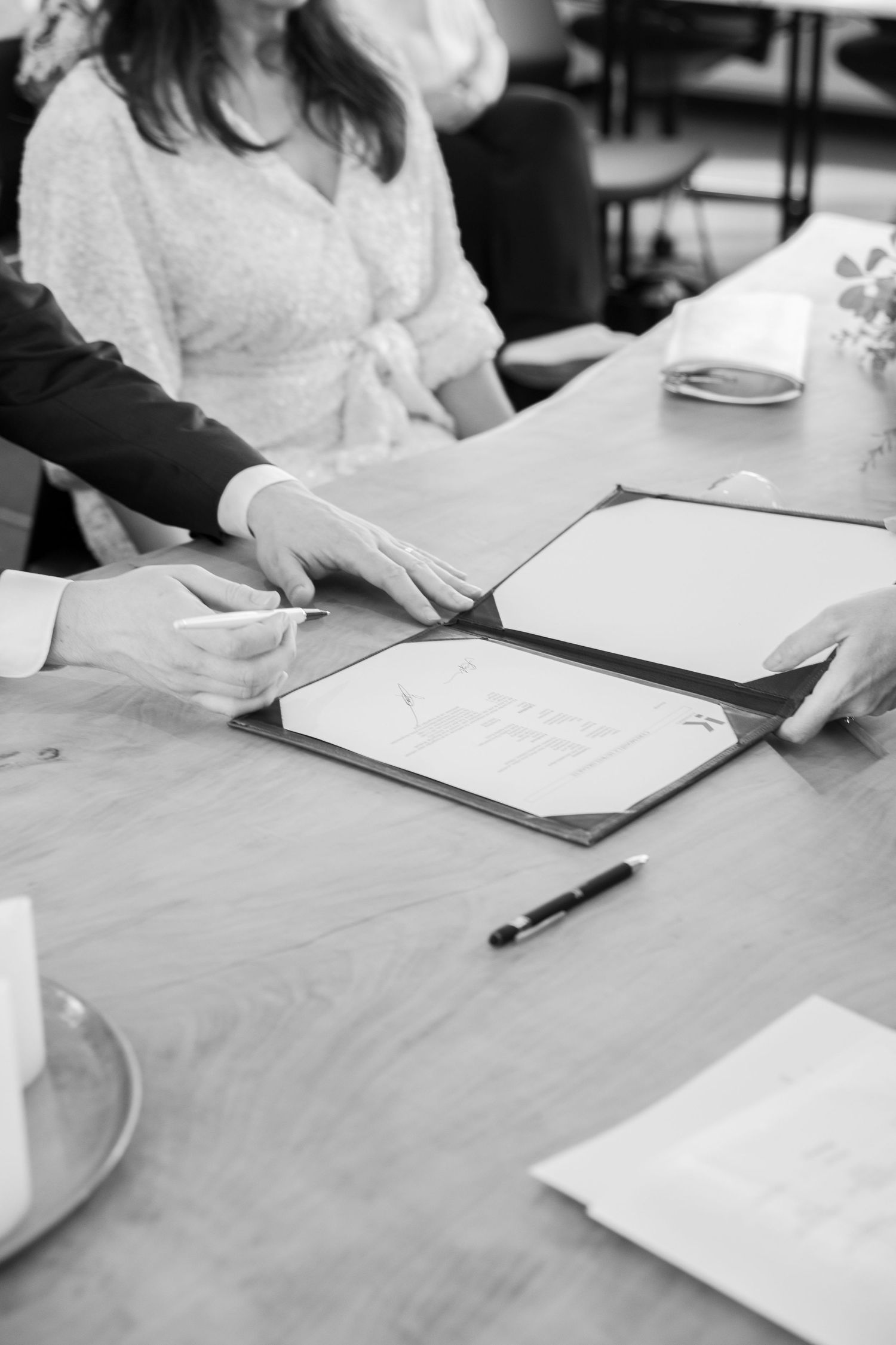 Black and white photo of hands signing wedding documents at a wooden table during a marriage ceremony.
