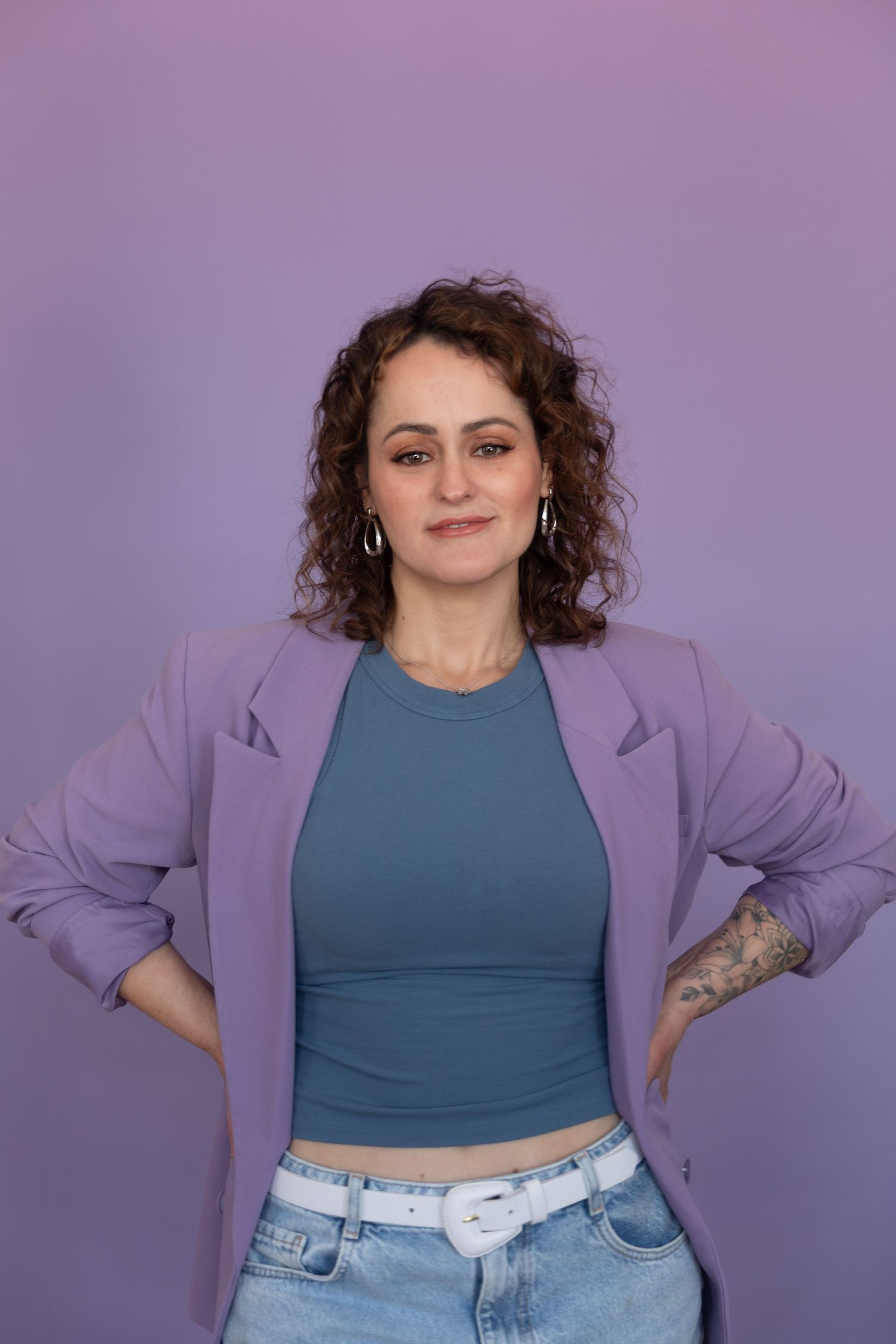 Woman with curly hair in purple blazer, teal crop top, and jeans posing confidently against purple background.