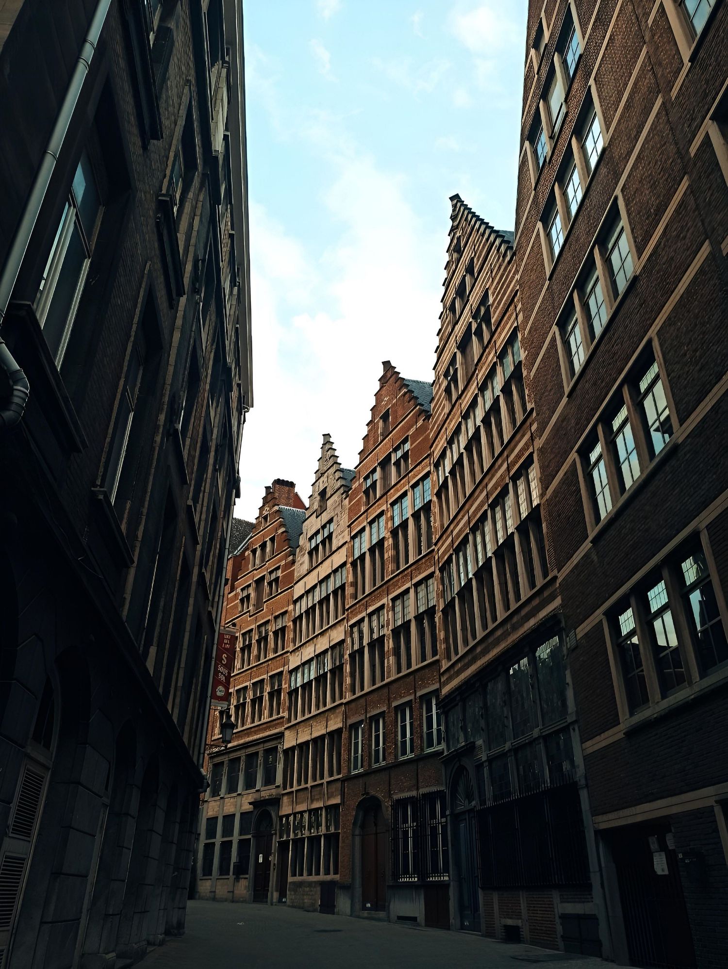 Narrow alley between historic red brick buildings with stepped gable rooflines in Antwerp, Belgium, under a cloudy sky.