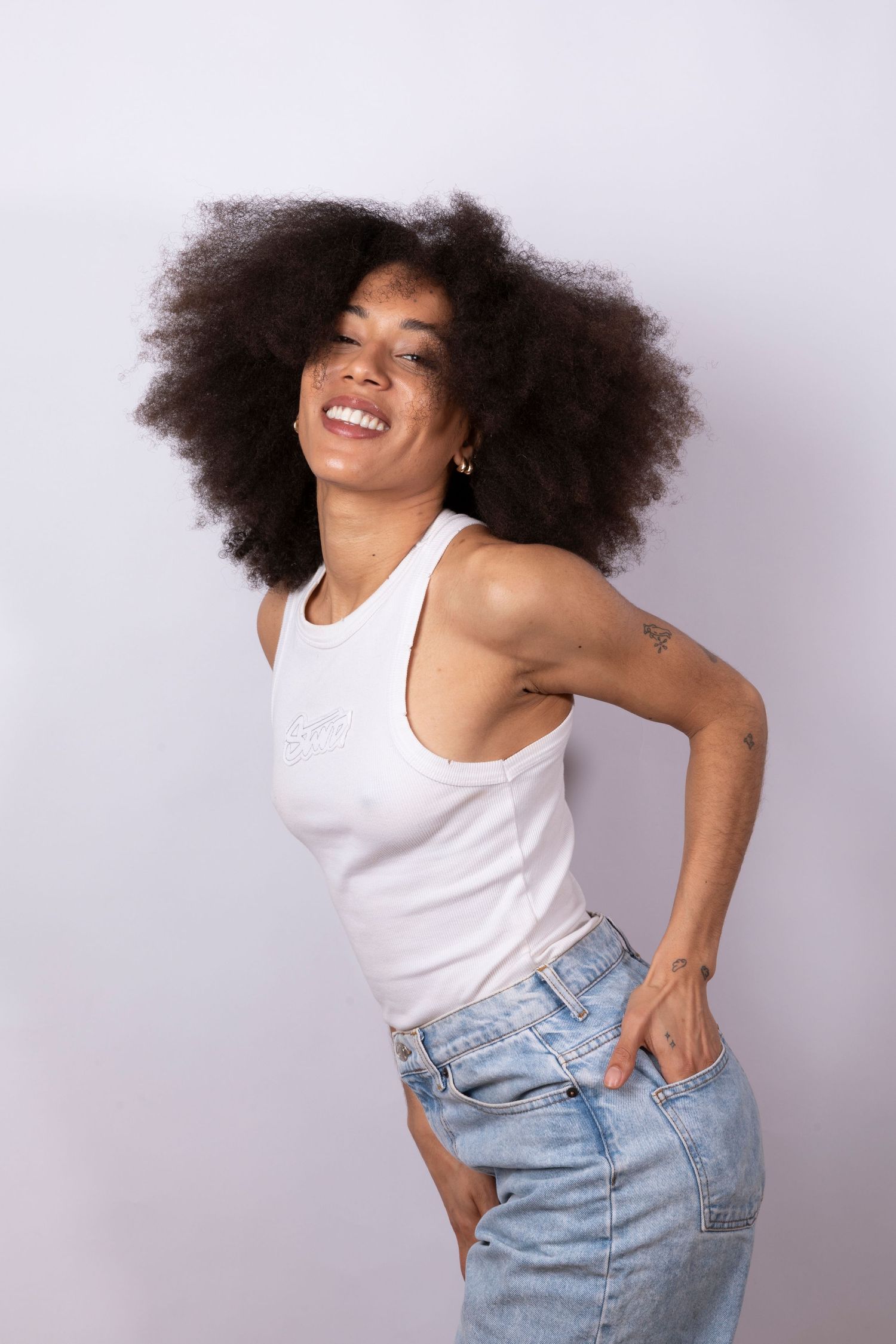 Smiling young woman with afro hair wearing white tank top and jeans posing confidently against light background.