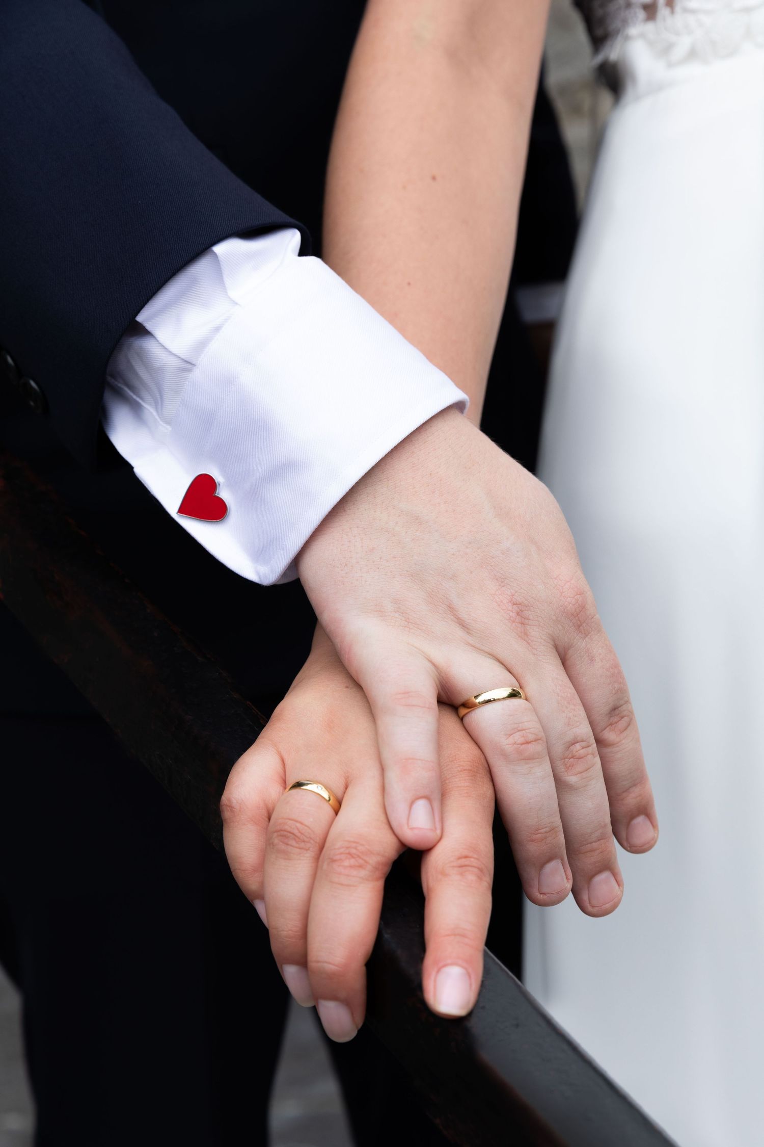 Newlywed couple holding hands showing gold wedding rings, groom wearing white shirt with red heart cufflink.