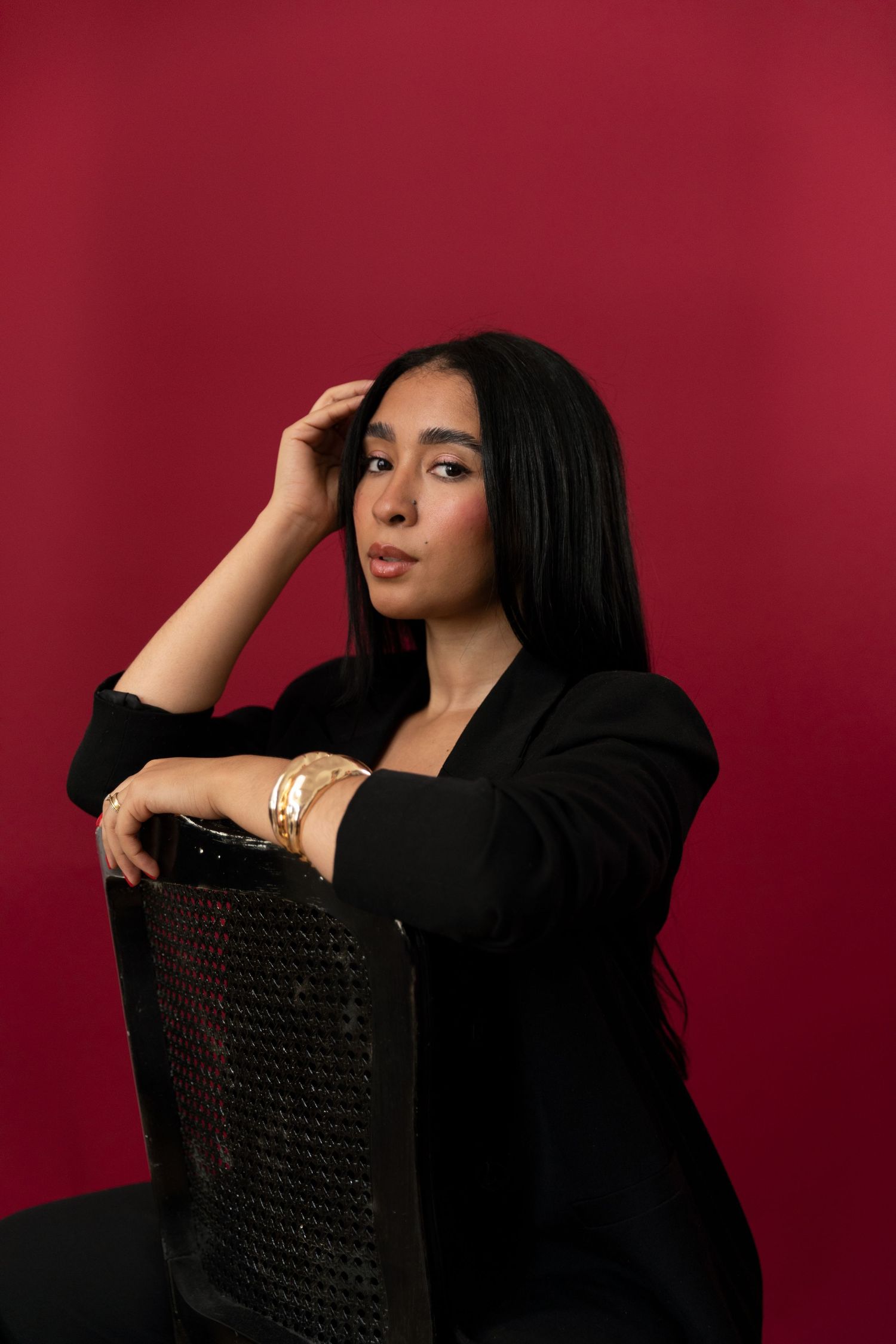 Young woman in black blazer with gold bracelet poses on chair against red background.