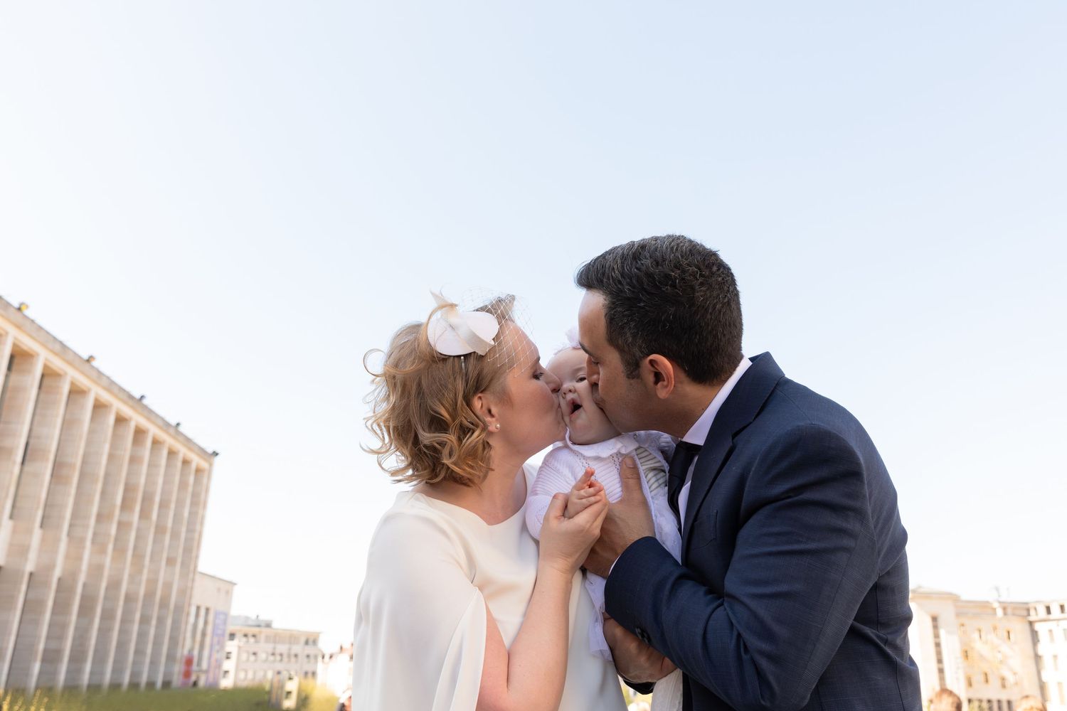 A couple in formal attire kisses their baby in white at an outdoor wedding ceremony.