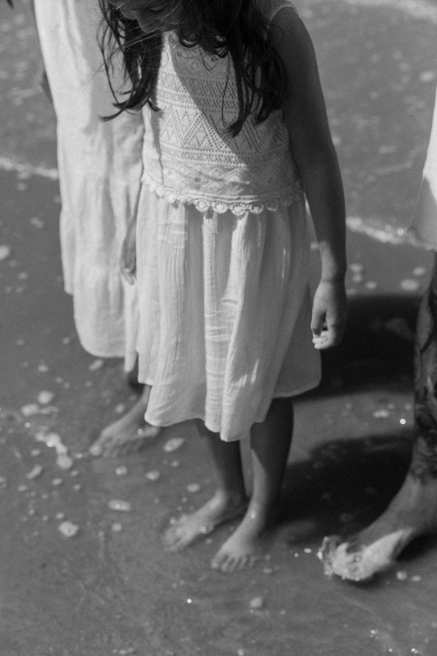 Black and white photo of a barefoot girl in a white lace dress standing on a sandy beach. photography brussels