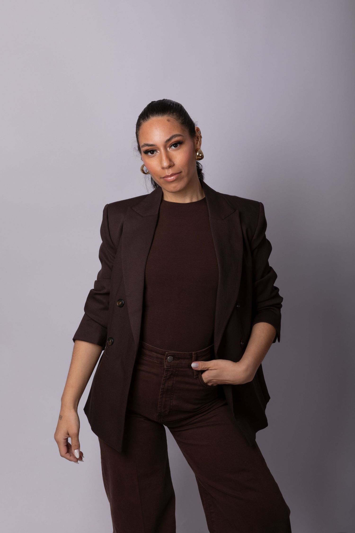 Woman in brown blazer, top, and pants poses confidently against a gray background.