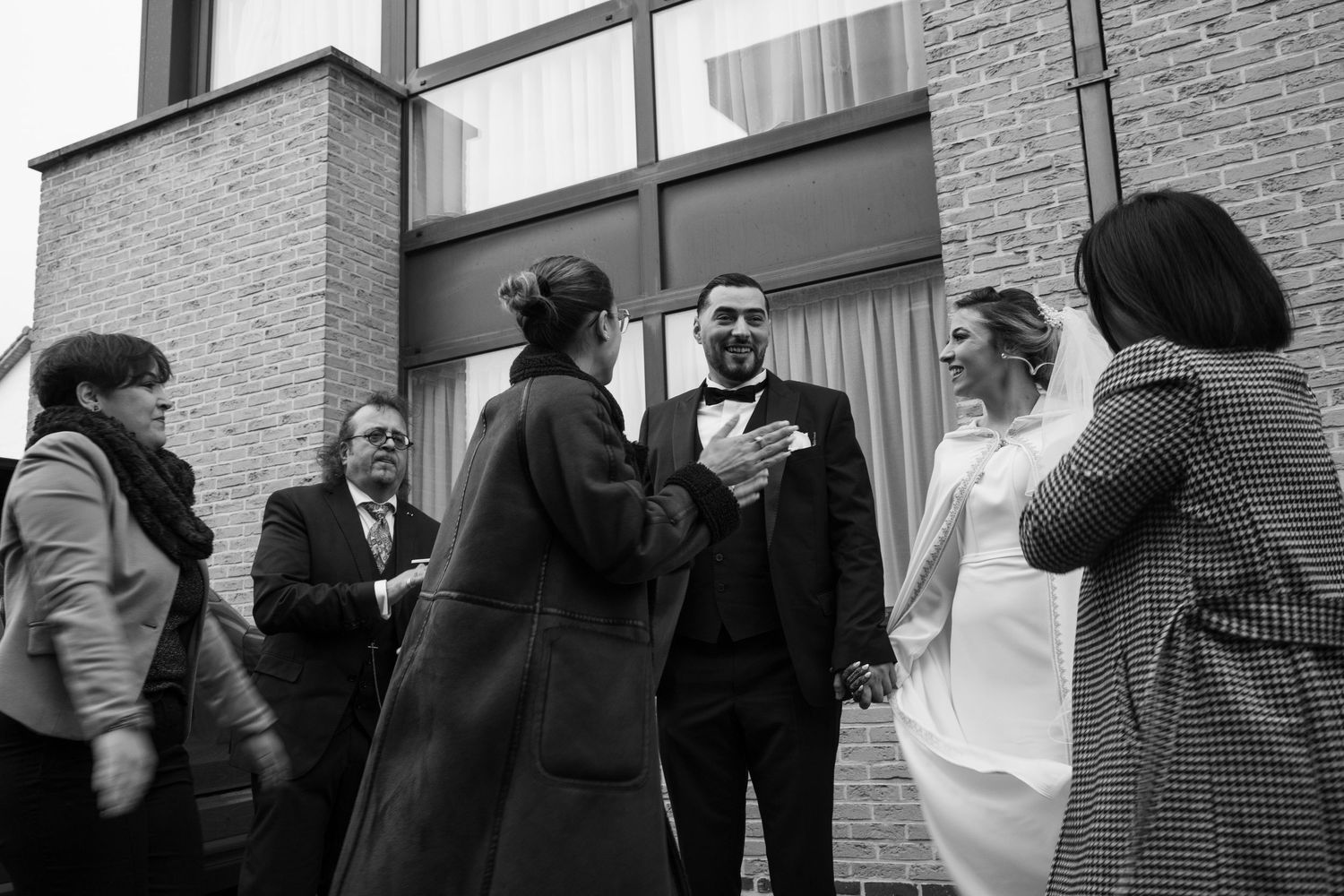 Black and white wedding photo of bride and groom greeting guests outside a brick building.