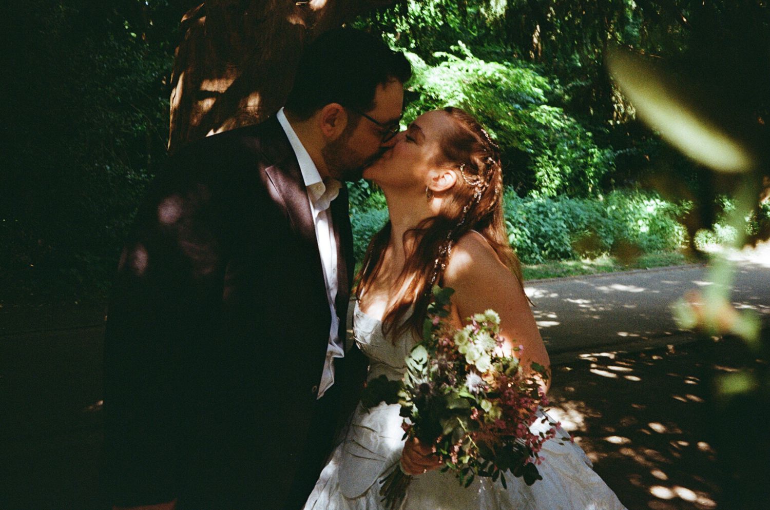 Bride and groom sharing a romantic kiss outdoors surrounded by lush green trees on their wedding day.