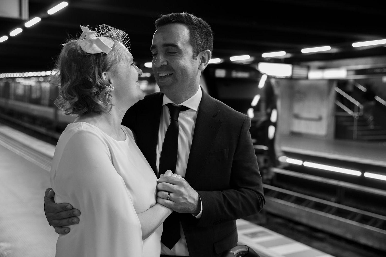 Black and white photo of a smiling couple in wedding attire sharing a tender moment on a subway platform.
