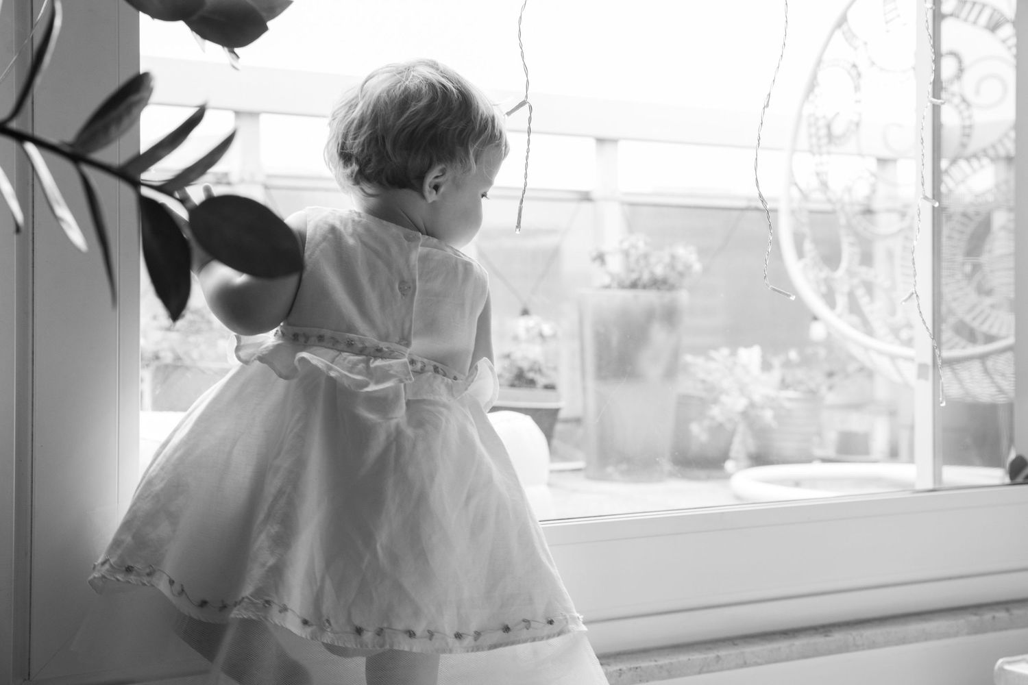 Black and white photo of a toddler girl in a white dress standing at a window looking outside.