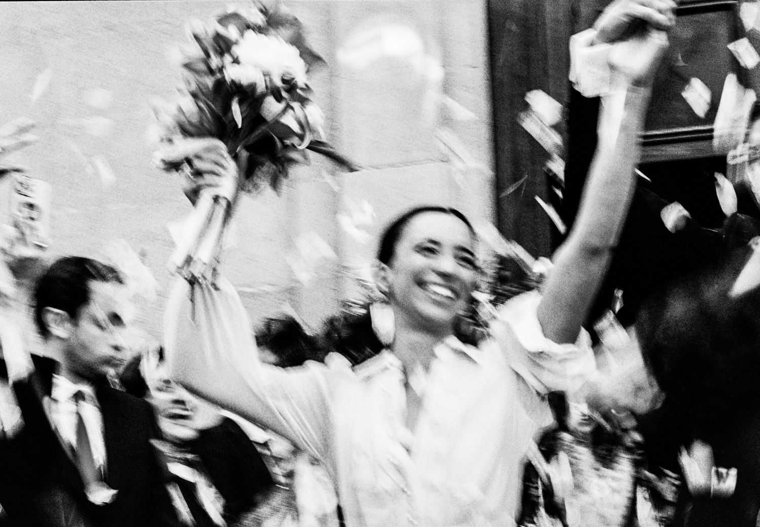 Black and white photo of a joyful bride raising bouquet amid celebration confetti.