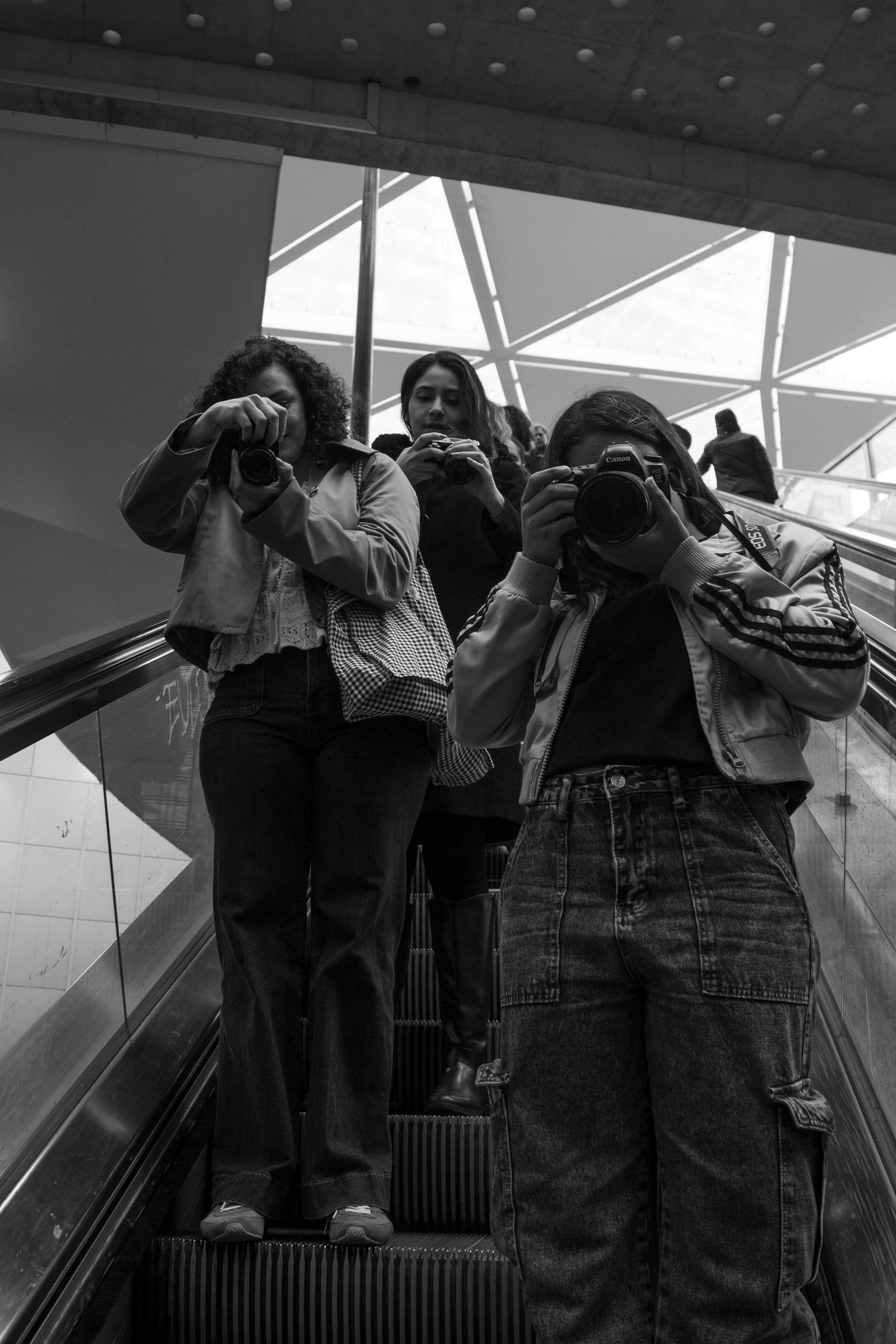 Three photographers on escalator holding cameras up, captured in black and white street photography style.