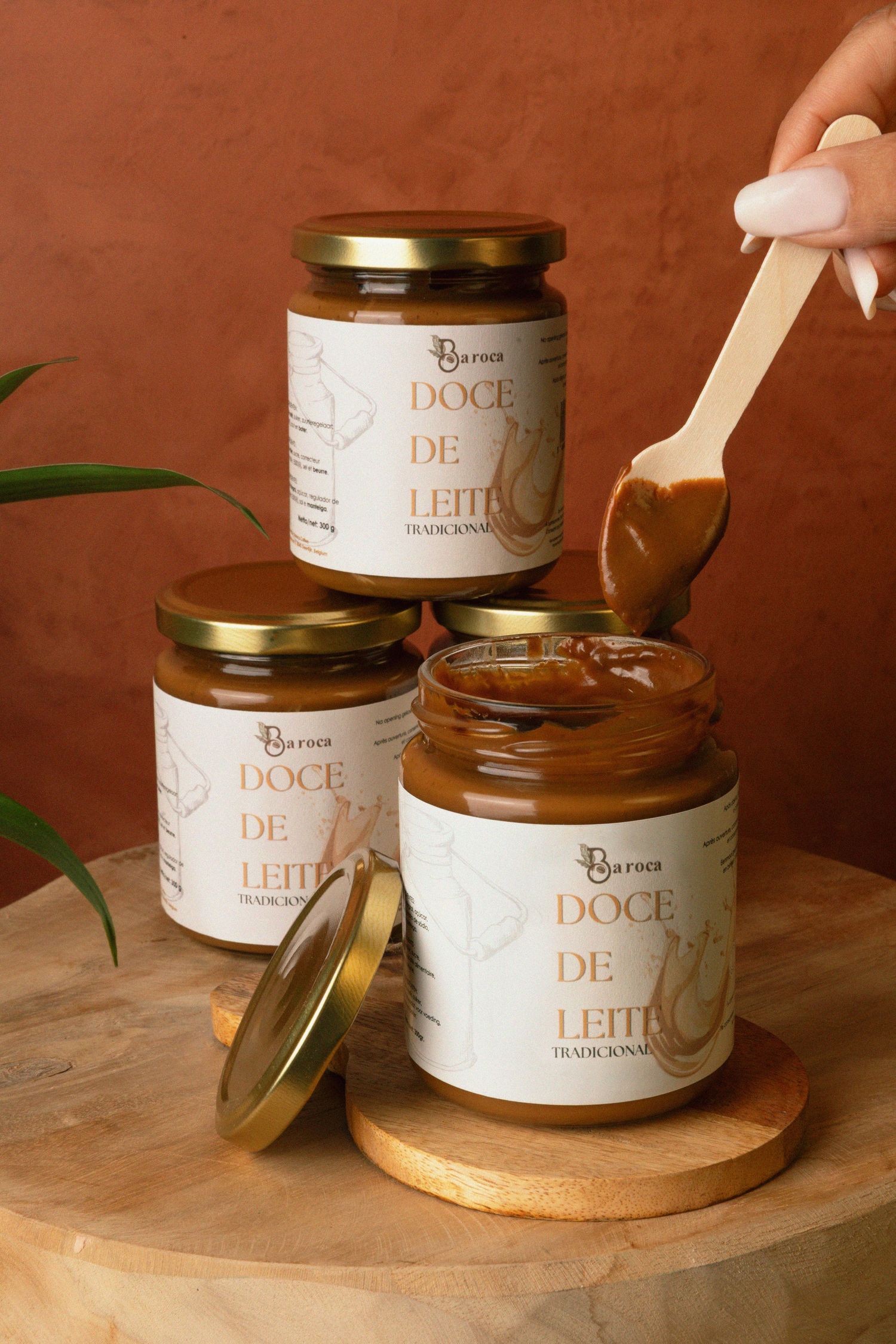 Four jars of Doce de Leite traditional caramel spread stacked on wood, with a hand holding a wooden spoon dripping caramel.