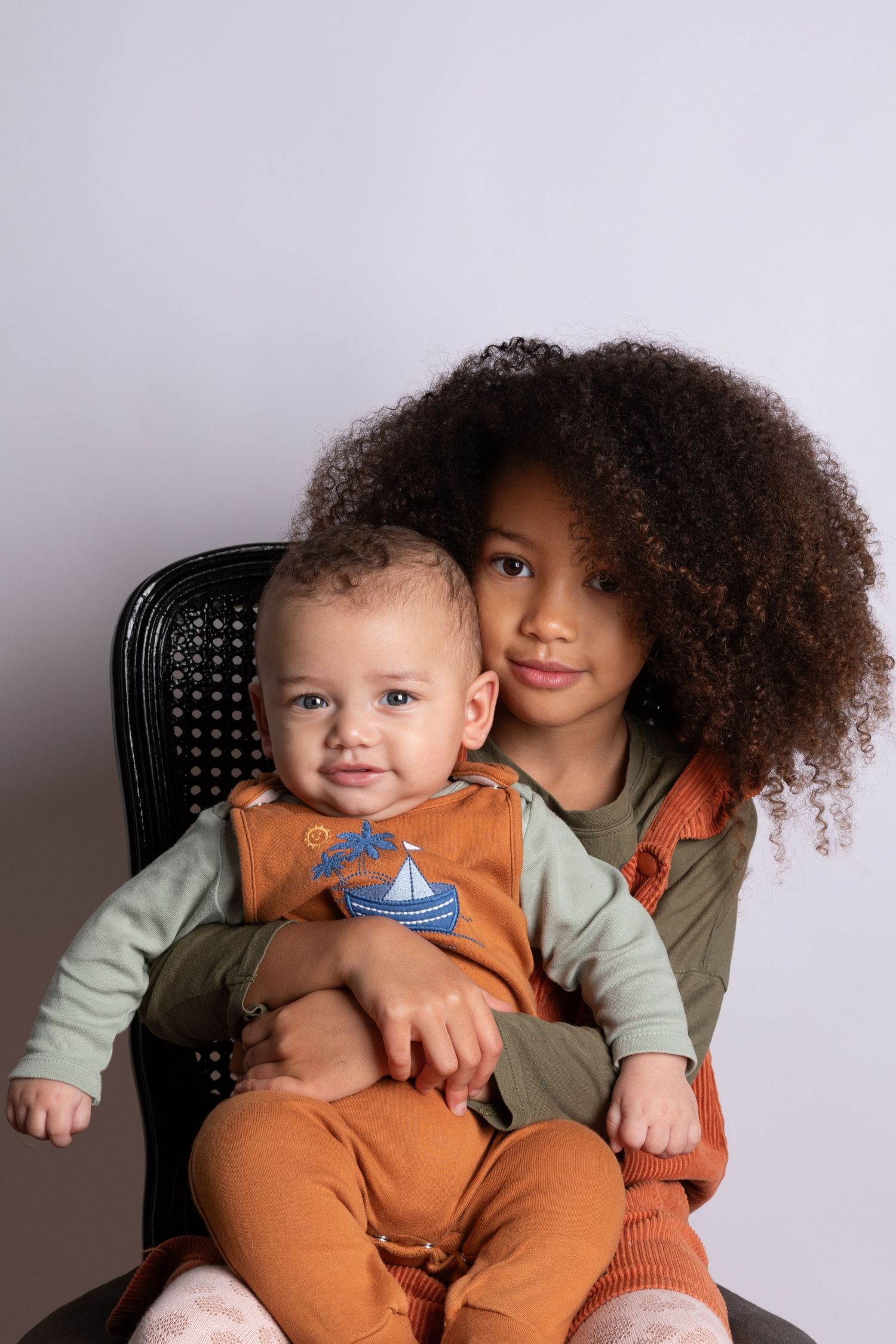 Young girl with curly hair holds a smiling baby in orange overalls while seated in a black chair.