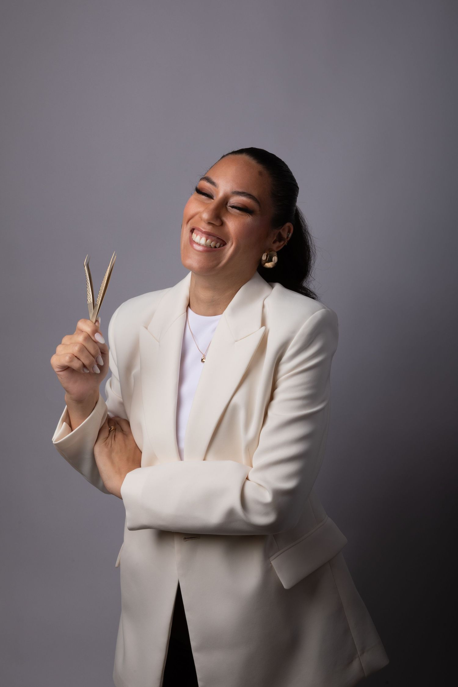 Smiling woman in white blazer holding gold scissors against gray background.