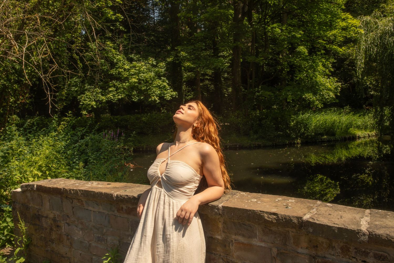 A woman in a white sundress with red hair tilts her head back in sunlight beside a stone wall in a lush green park.