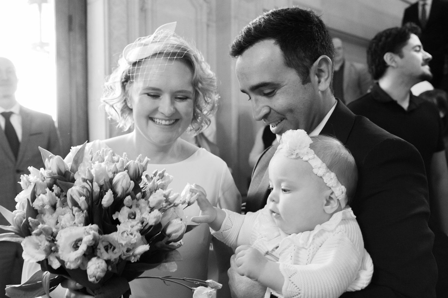 Black and white wedding photo of smiling bride with bouquet and groom holding baby in white outfit.