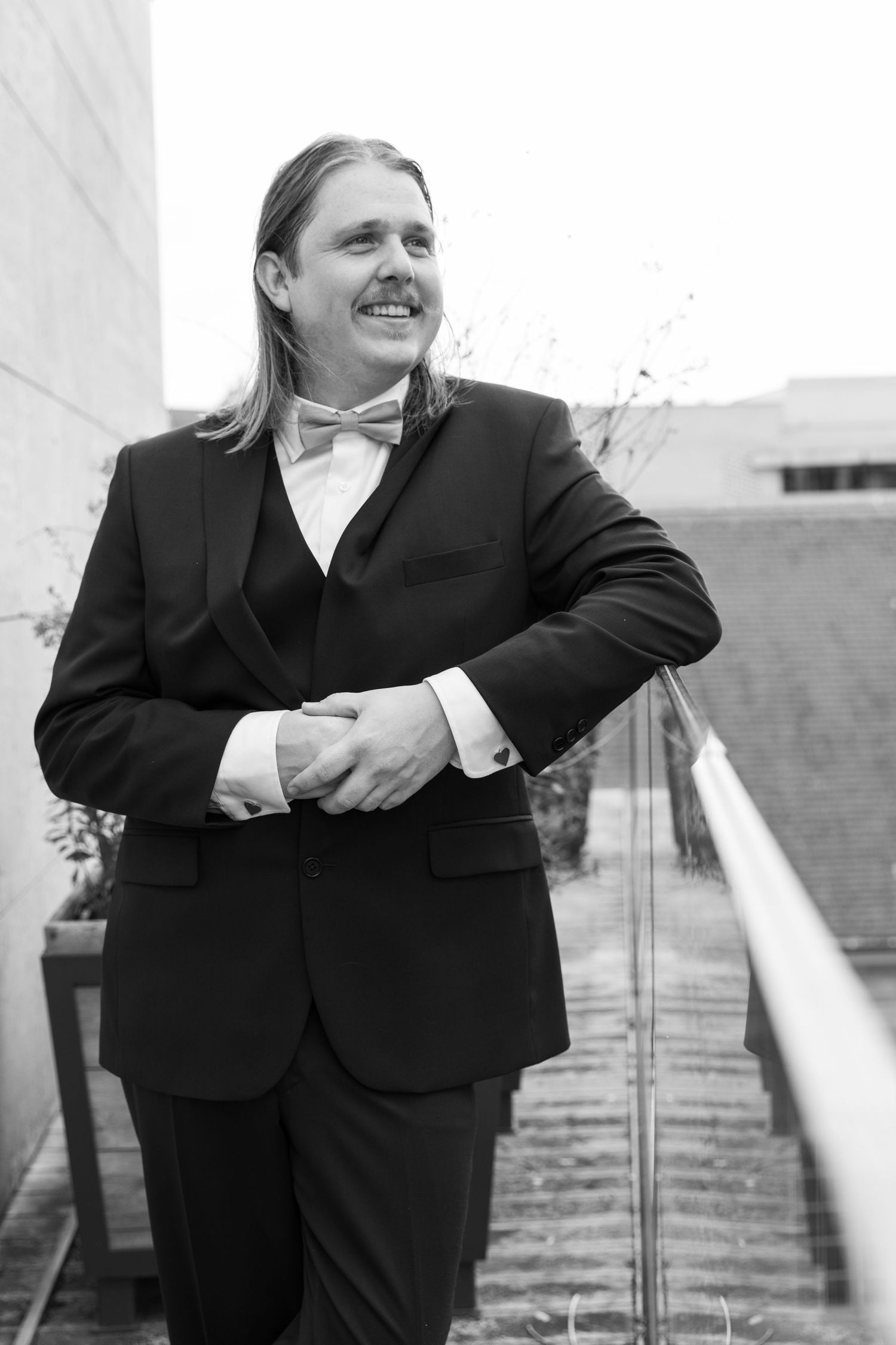 Elegant groom in black tuxedo with bow tie, long hair and mustache, leaning on glass railing outdoors.