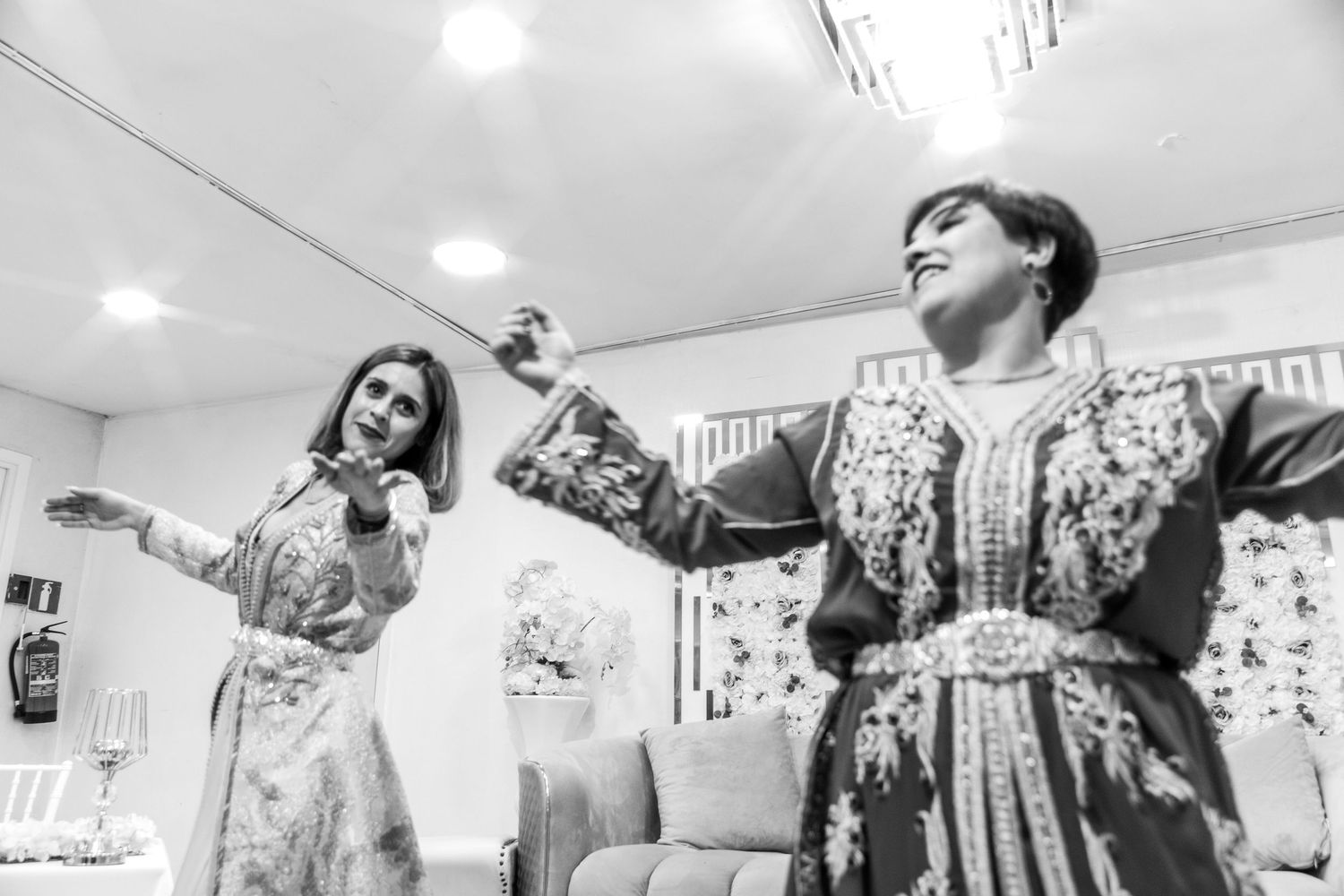 Two women in ornate traditional Moroccan caftans dancing joyfully at a celebration event.